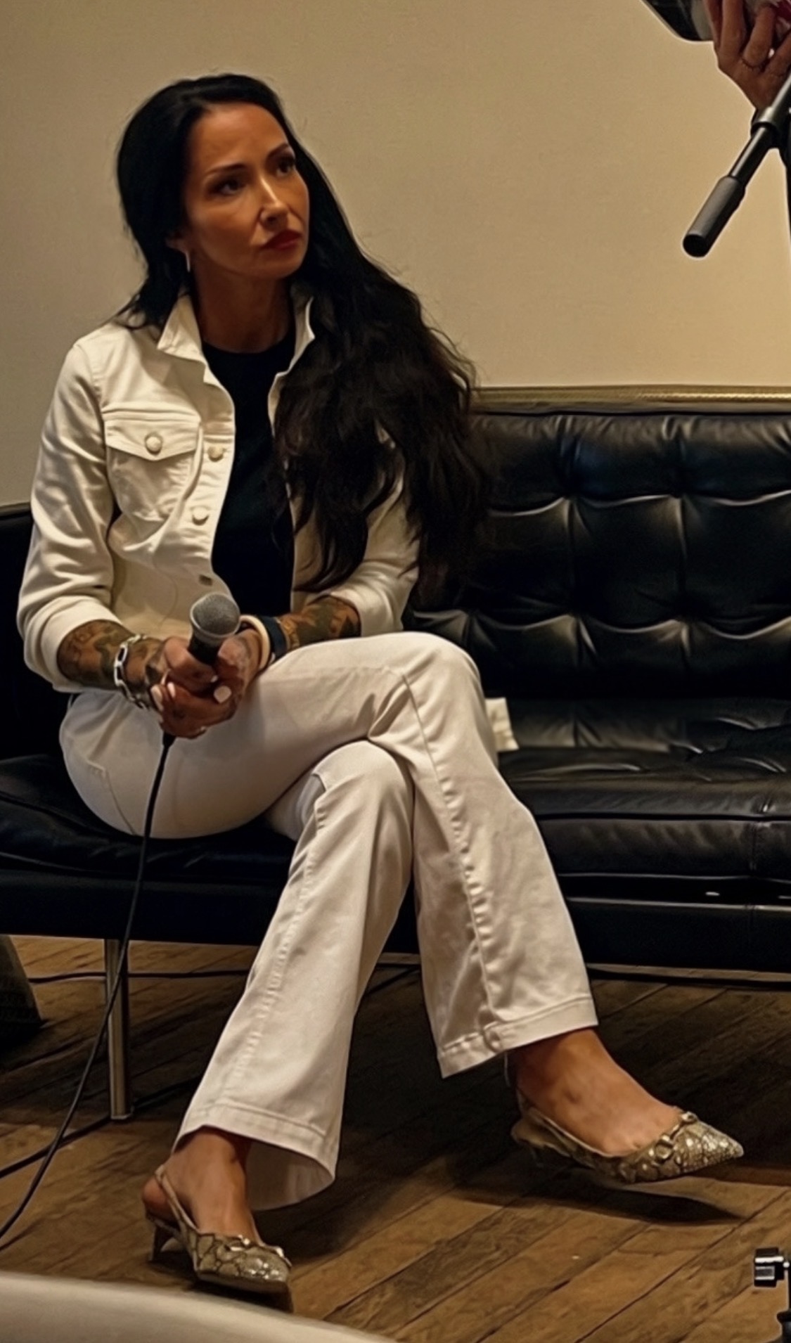 Woman with long dark hair sitting on a black leather couch, holding a microphone, wearing a white outfit and glittery shoes.
