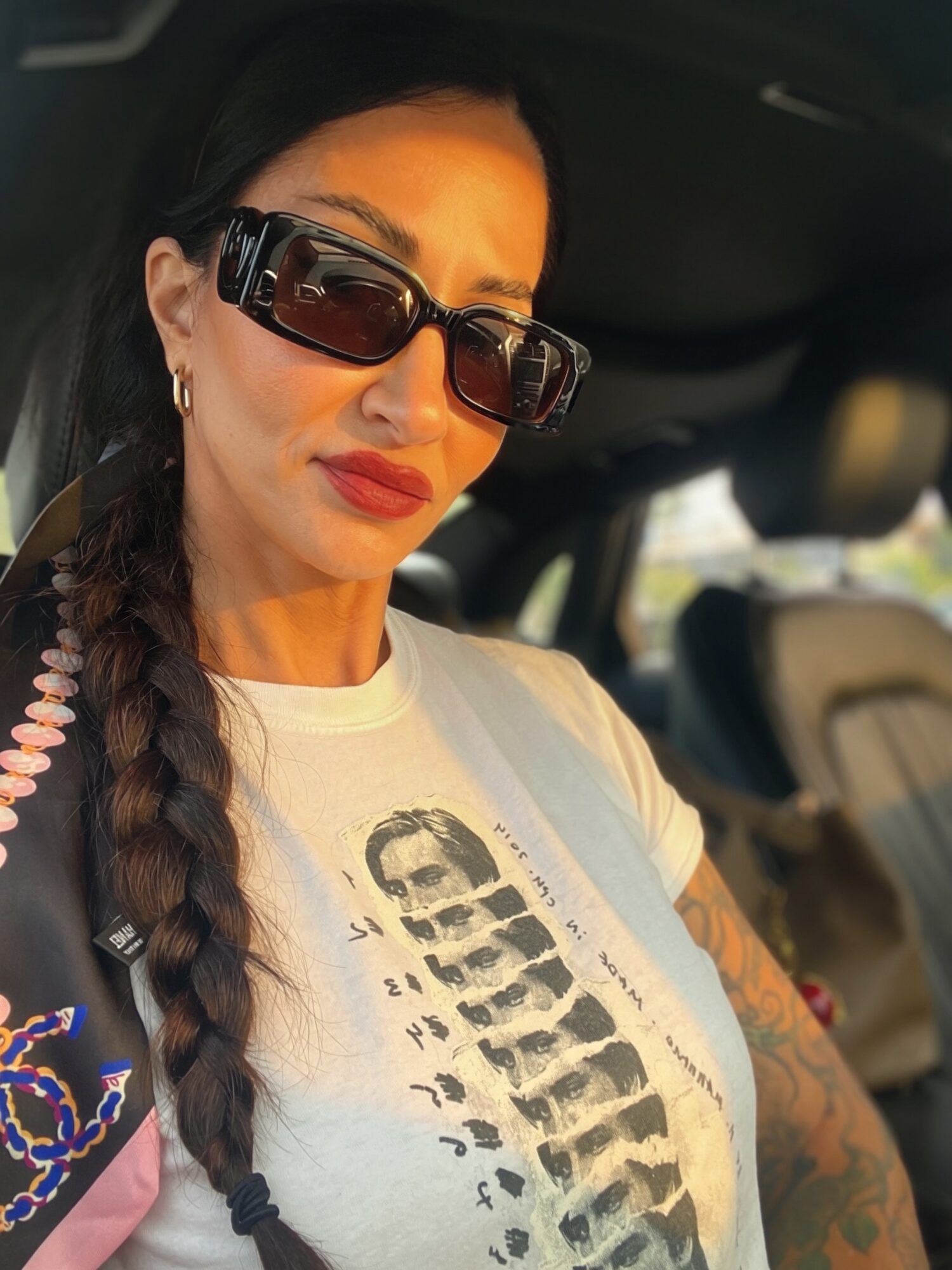 Woman with long braid, sunglasses, and red lipstick sitting in a car, wearing a graphic t-shirt, looking at the camera.