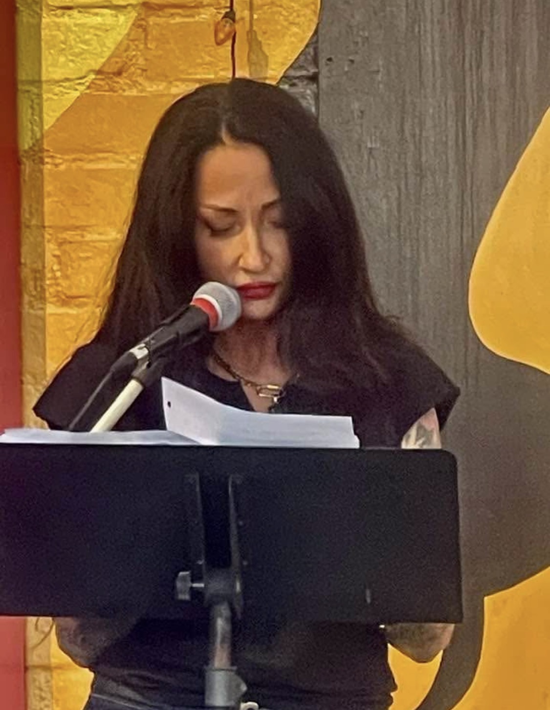 Woman with long dark hair reading from paper at microphone, in front of colorful background.