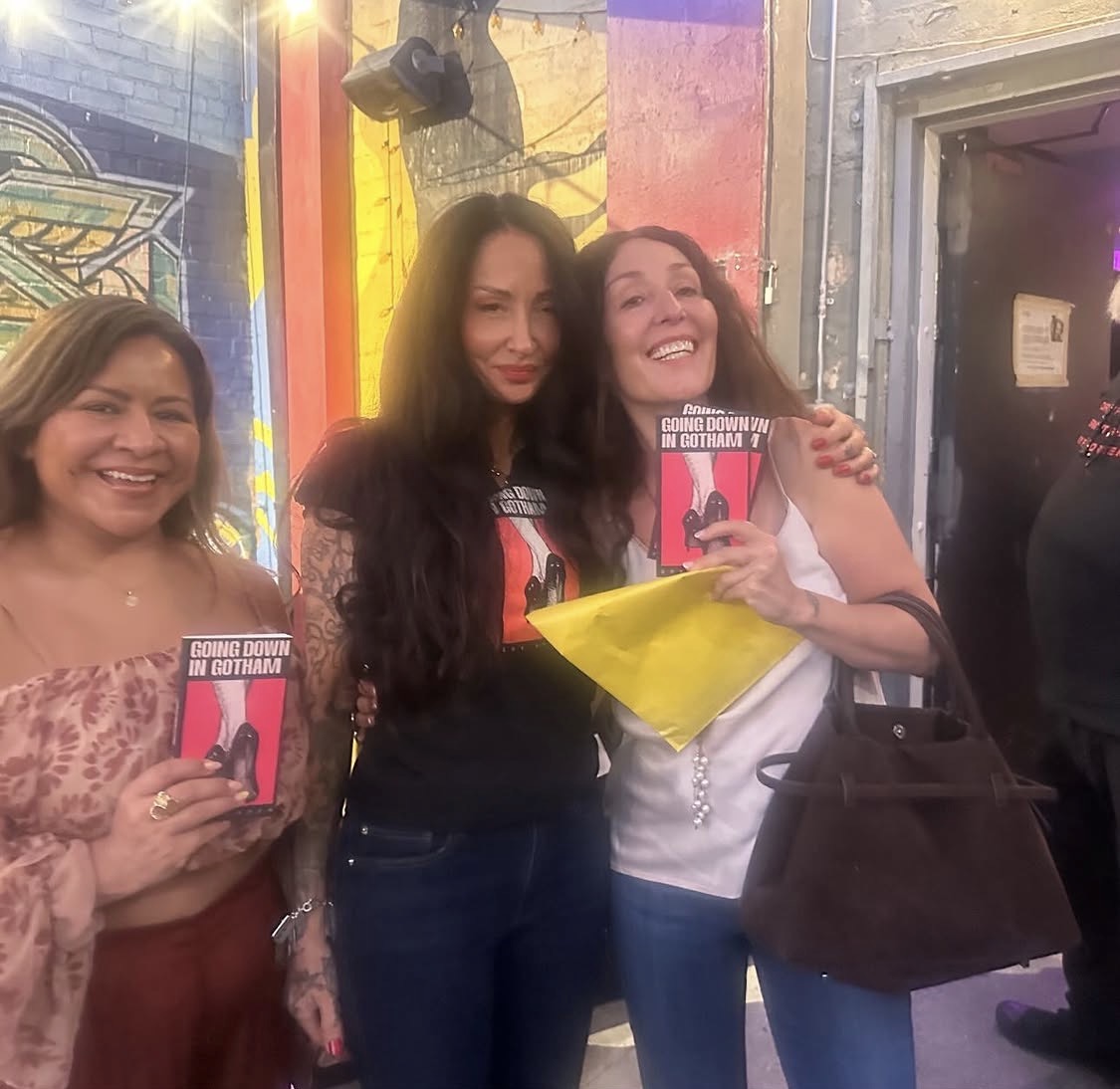 Three women smiling, two holding books, standing indoors with colorful background, one woman hugging another, all appear happy.