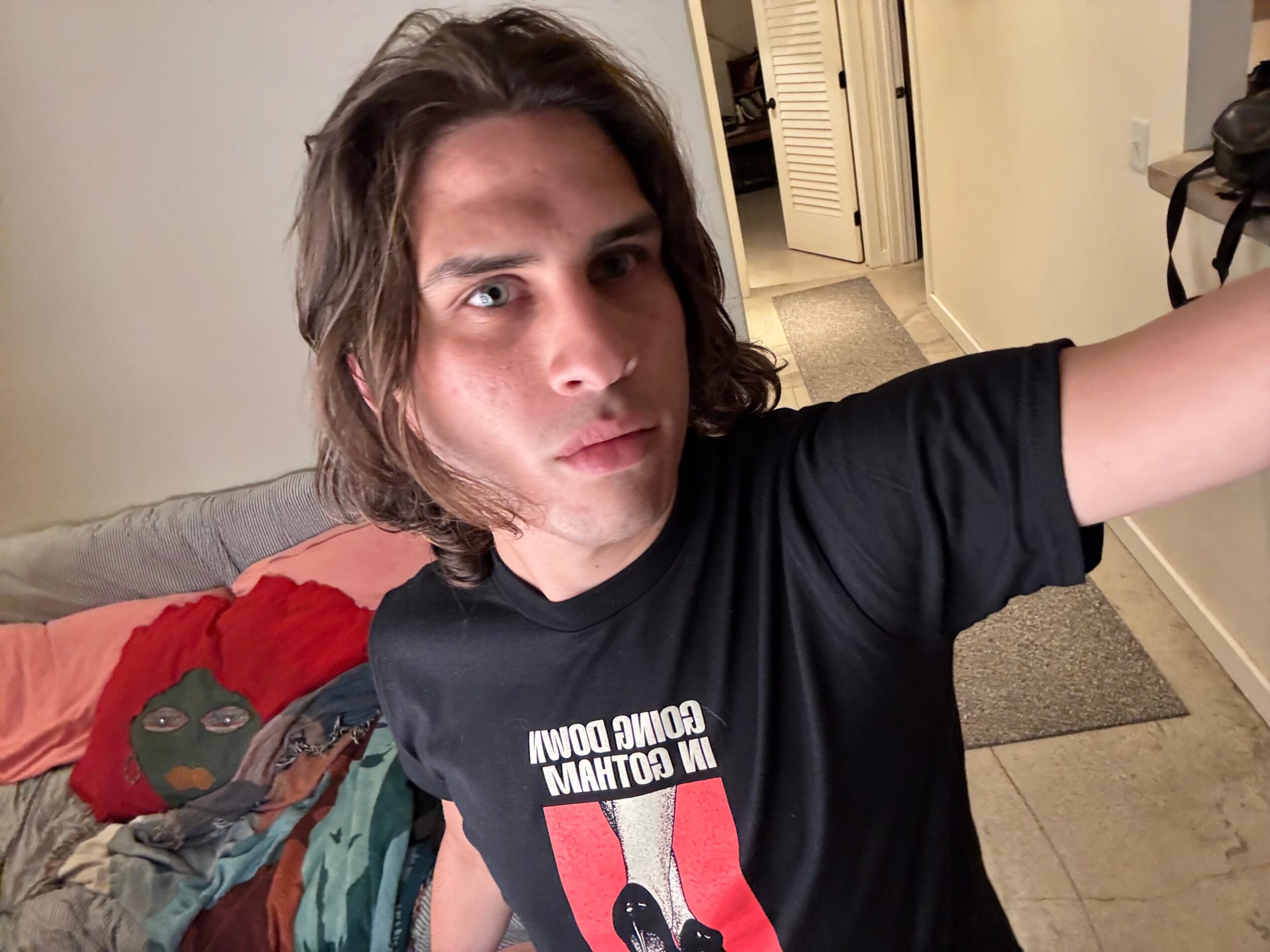 Person with shoulder-length hair taking a selfie indoors, wearing a black T-shirt with text and a graphic, standing near a doorway.
