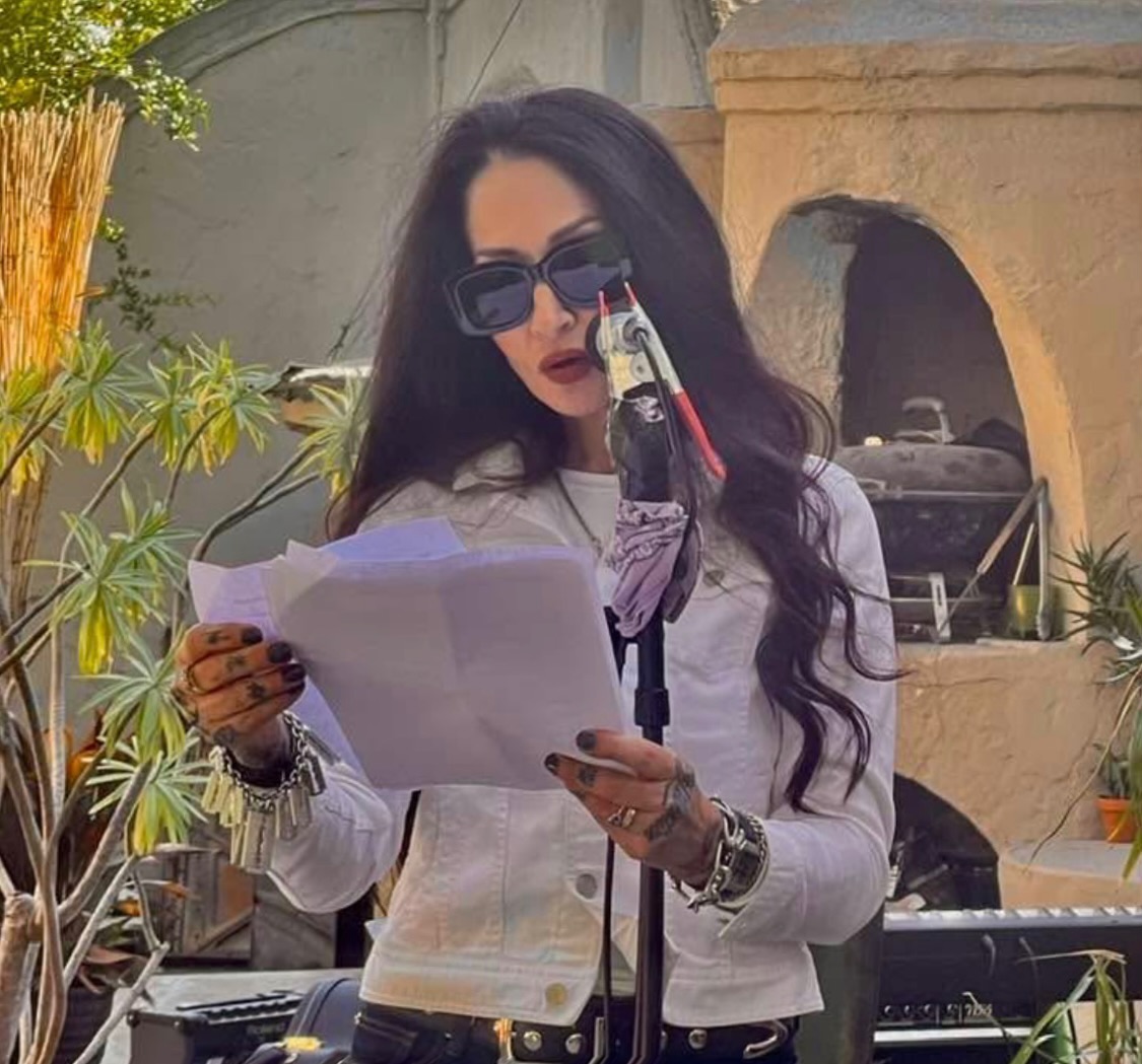 Woman with long dark hair, sunglasses, and white jacket reading paper at outdoor event, with equipment and plants in background.