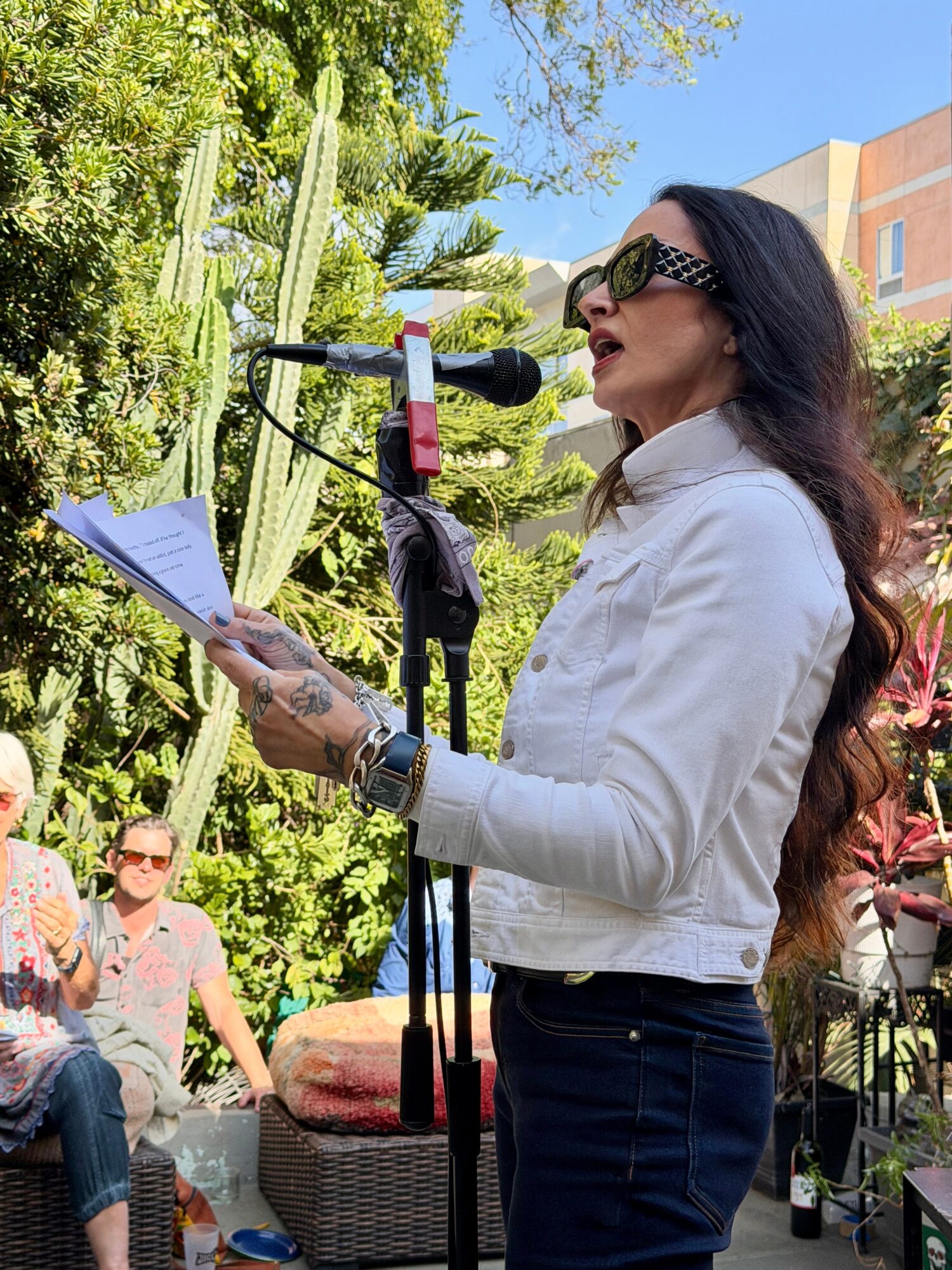 Woman with long dark hair, sunglasses, white jacket, reading from paper at outdoor event, microphone in front, trees and people in background.