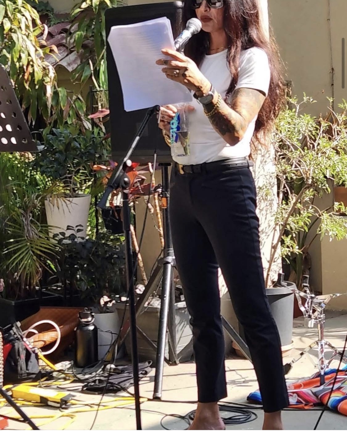 Woman with long hair, sunglasses, white t-shirt, and black pants reading from paper at outdoor event, surrounded by plants and equipment.