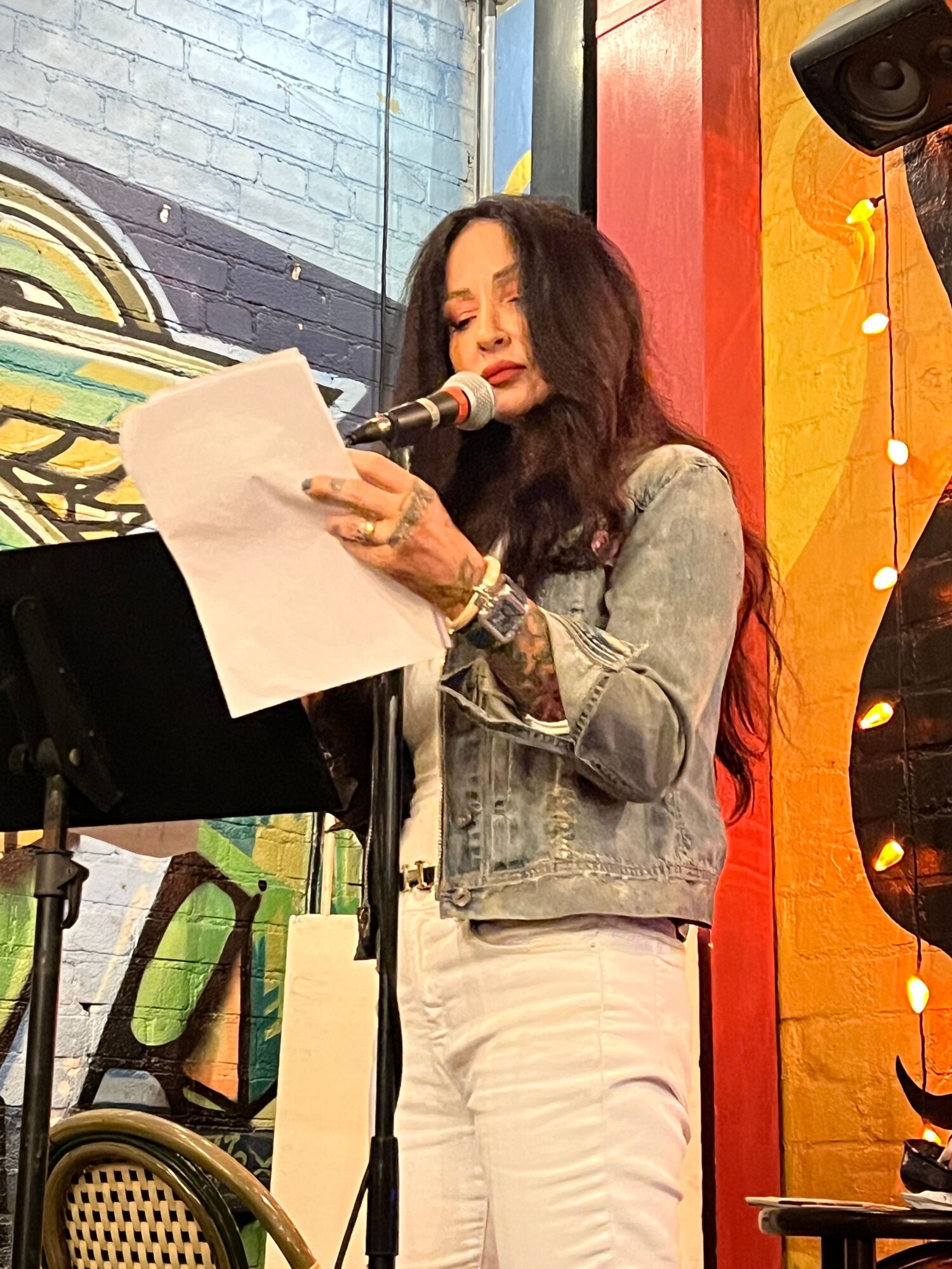 Woman with long dark hair, wearing a denim jacket and white pants, speaking into a microphone while holding papers.
