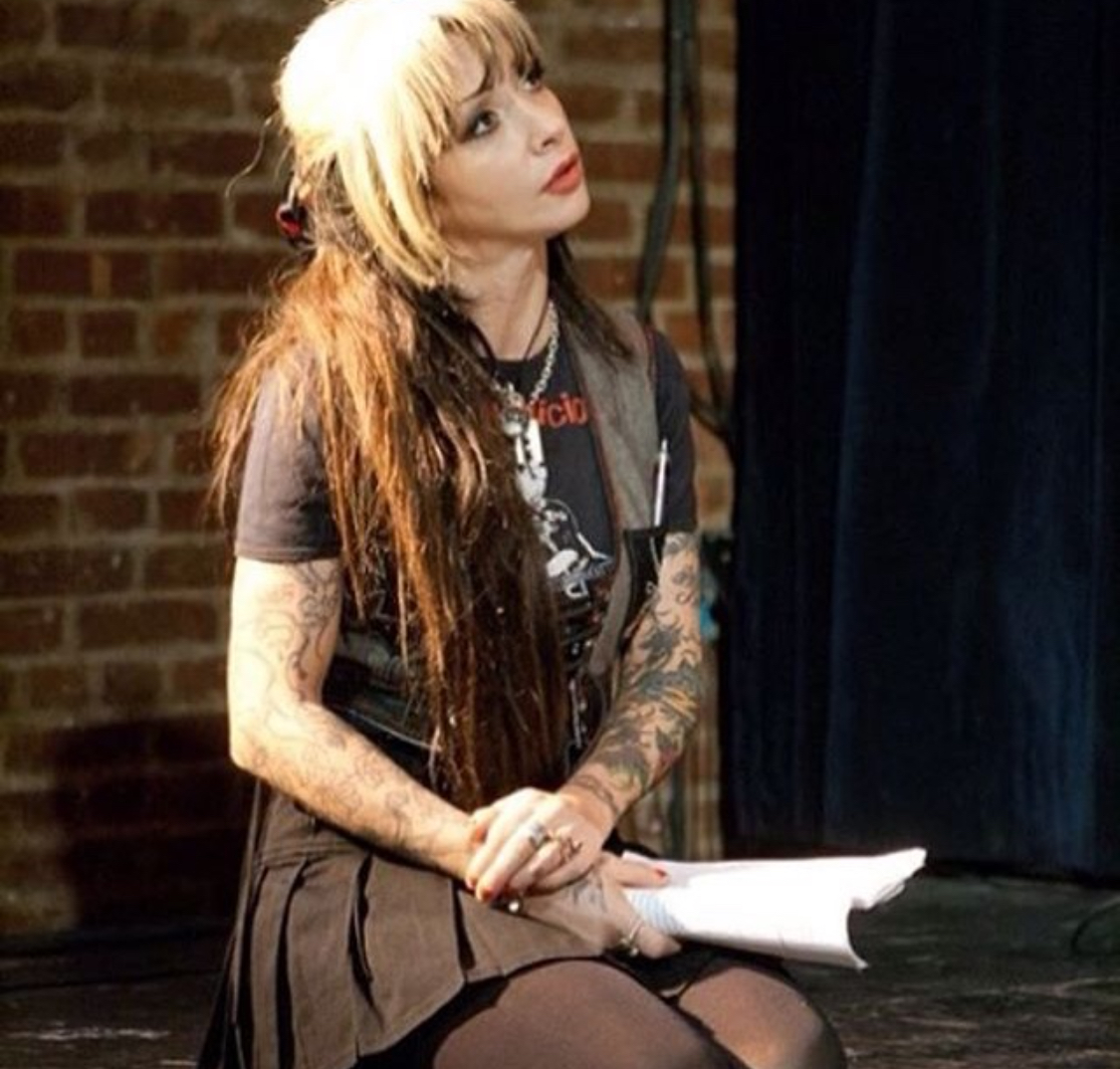 Young woman with long hair and tattoos sitting on stage holding papers, brick wall background.