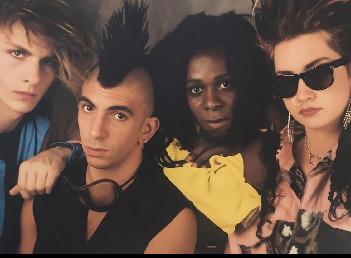 Four people pose closely, with diverse hairstyles and clothing, some wearing sunglasses, in a dark setting.