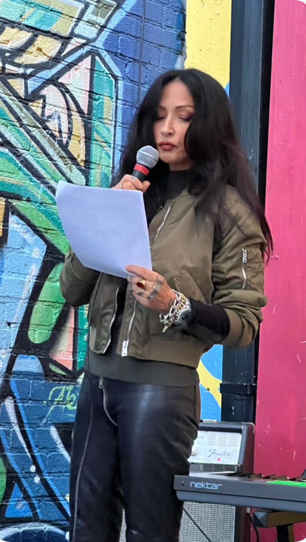 Woman with long dark hair reading from paper and holding a microphone, standing in front of colorful graffiti wall.