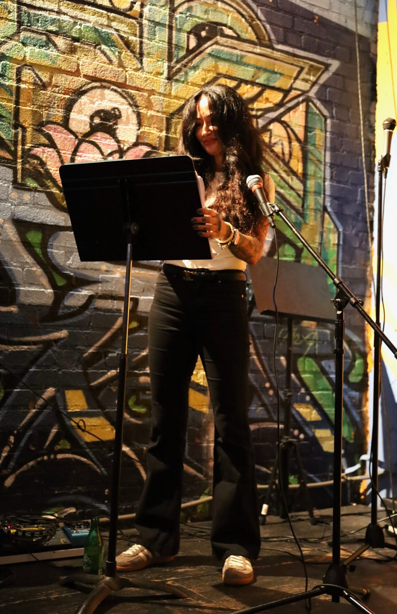 Woman standing on stage with microphone, reading from a music stand, graffiti wall background, stage equipment visible