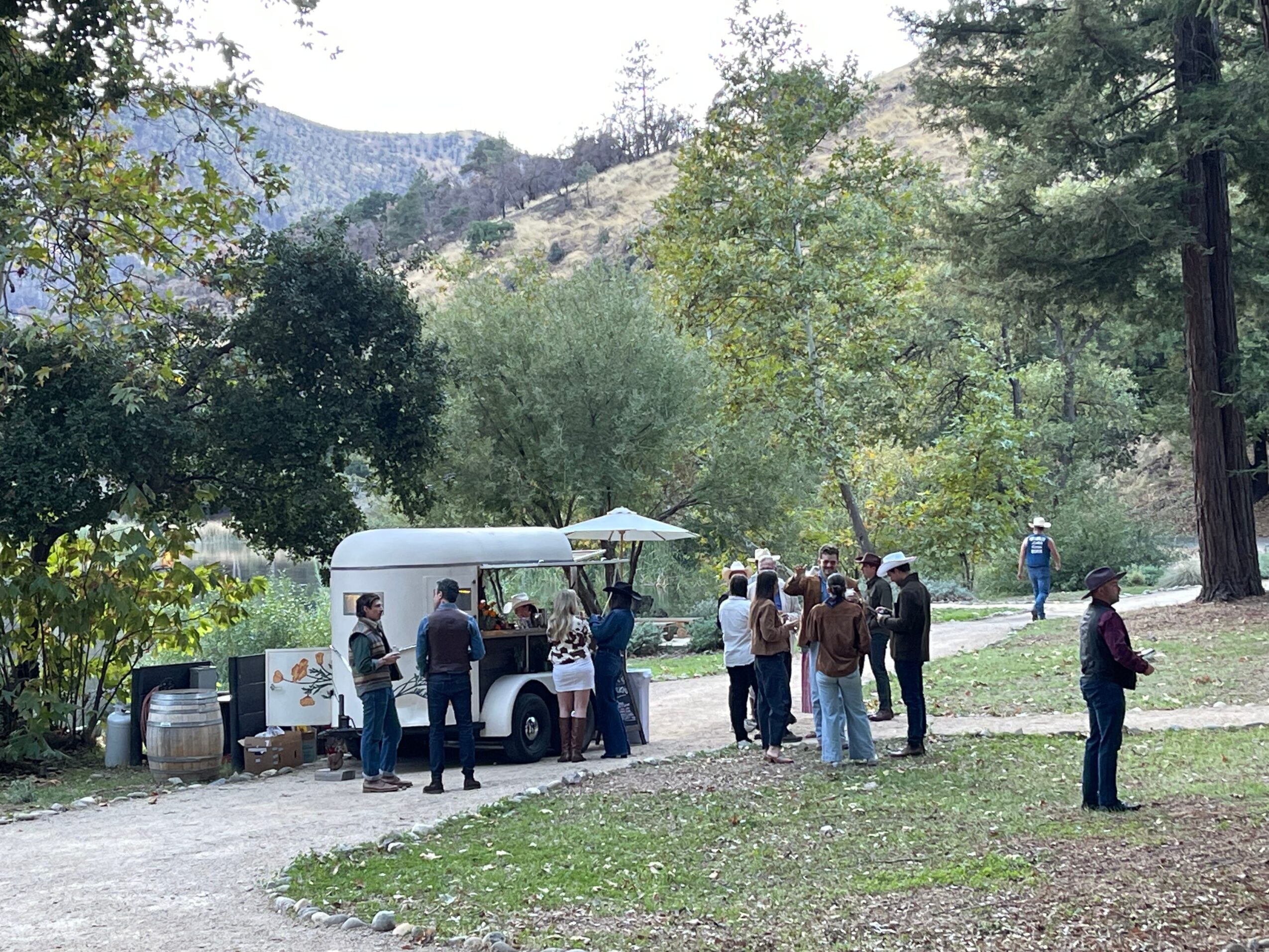 Group of people gathered around a food truck in a park with trees and mountains in the background.