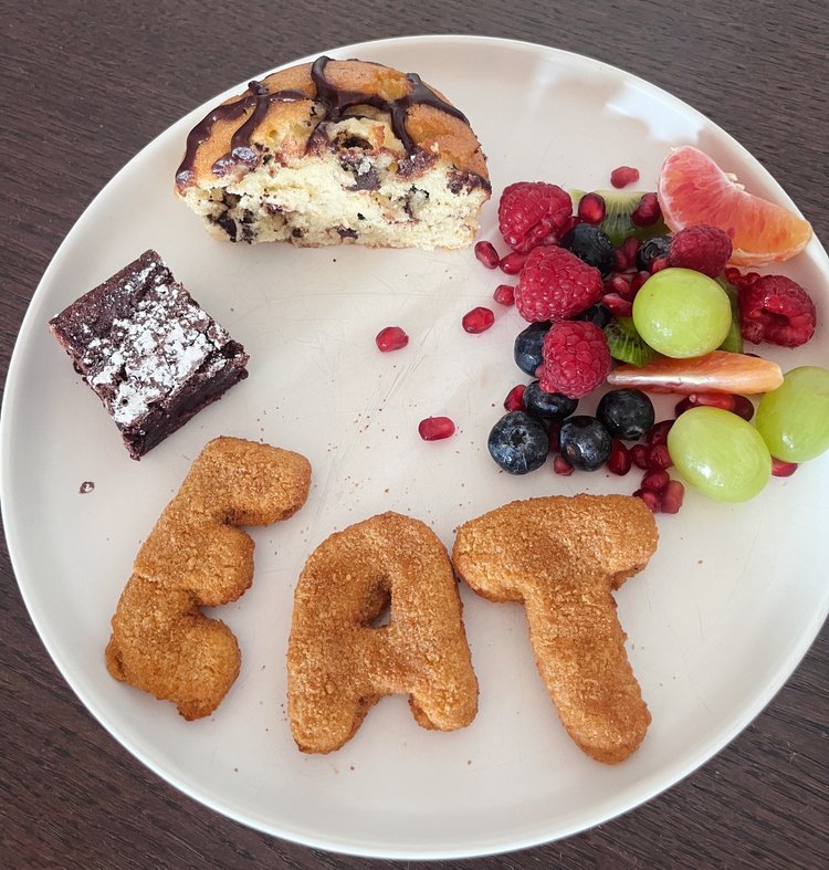 Plate with a slice of cake, brownie, fruit, and cookies spelling 'EAT'.