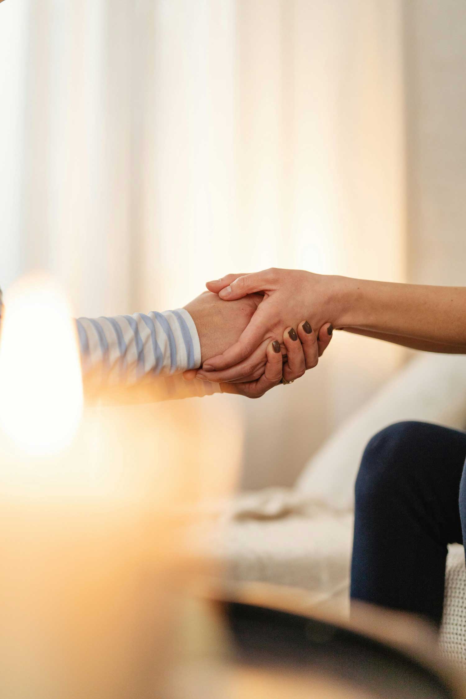 Two people hold hands, one with a striped sleeve, in a softly lit room with curtains in the background.