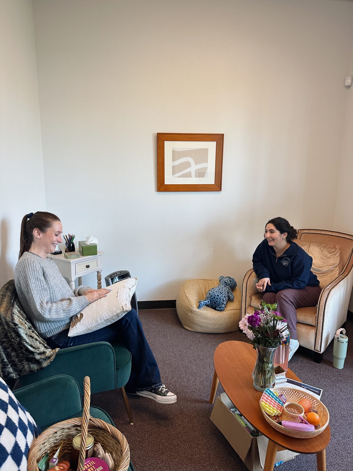 Two women sit in a living room, one on a teal chair and the other on a bed, with a small table and basket nearby.