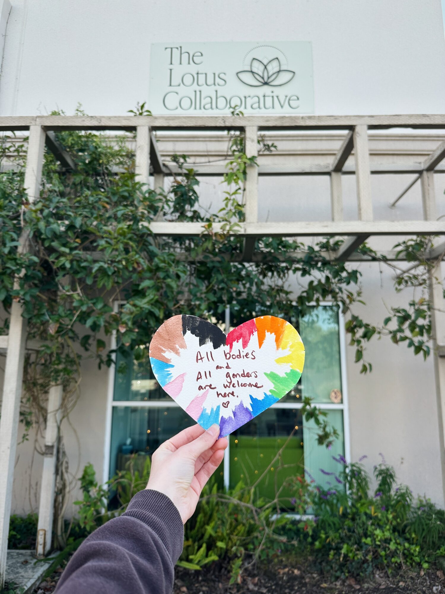 Person holding a colorful heart-shaped sign in front of a building with a sign reading 'The Lotus Collaborative'.