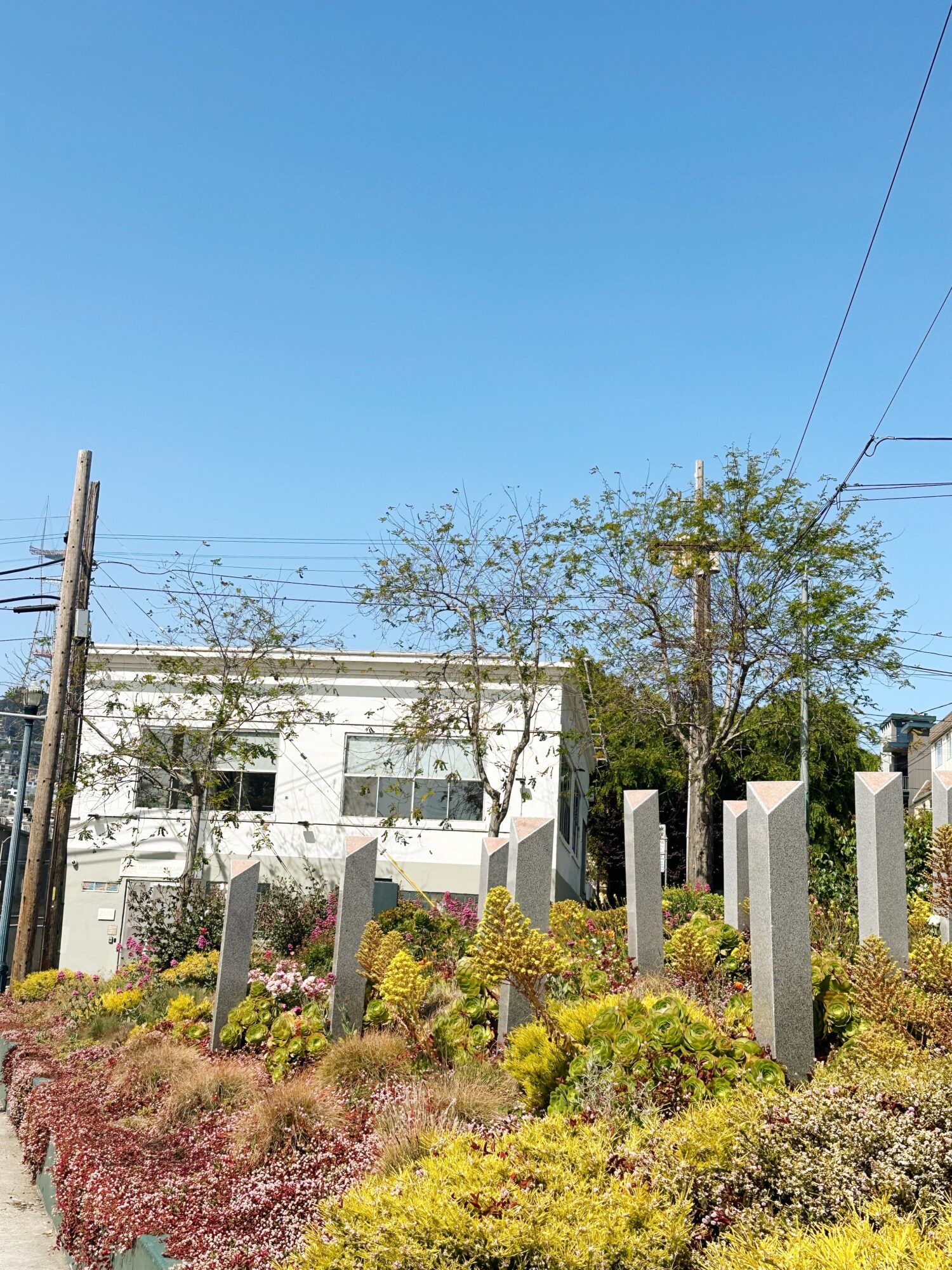 White building behind trees and a colorful garden with stone pillars, under a clear blue sky.