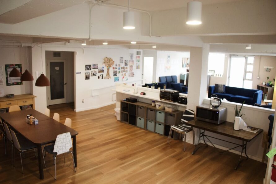 Open-plan room with wooden floor, dining table, chairs, and a lounge area with blue sofas and large windows.