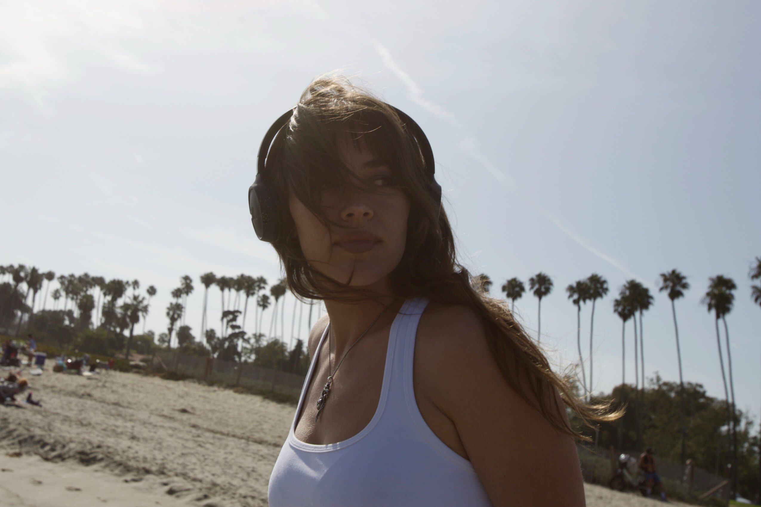 Woman with long hair wearing a white tank top at the beach, palm trees in background, sunlight overhead.