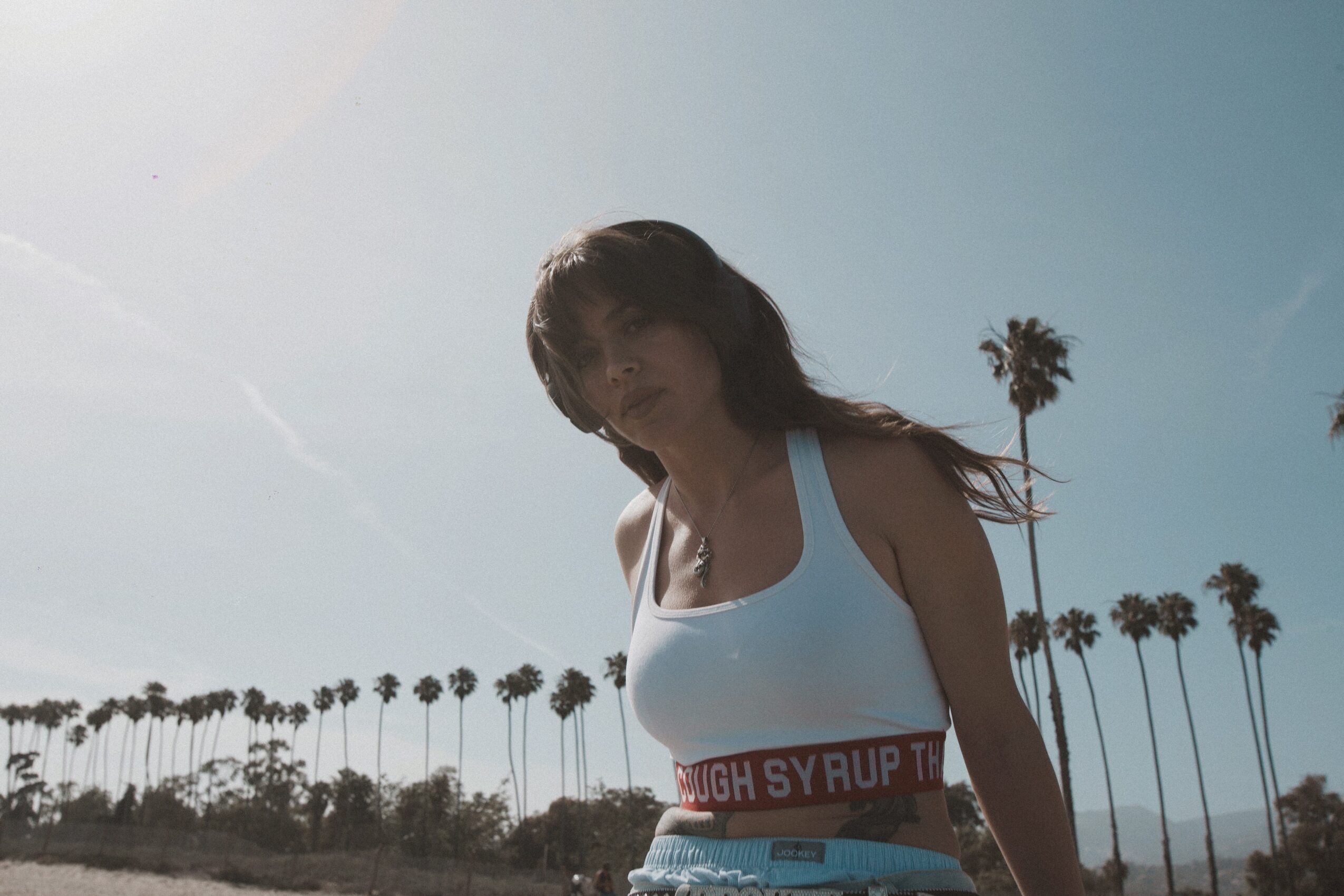 A woman with long hair in a white tank top stands outdoors with palm trees in the background under a clear sky.