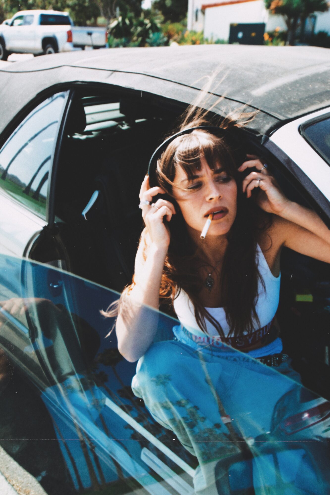 Young woman with curly hair wearing headphones, sitting in a car, looking at the camera, with sunlight shining on her face.