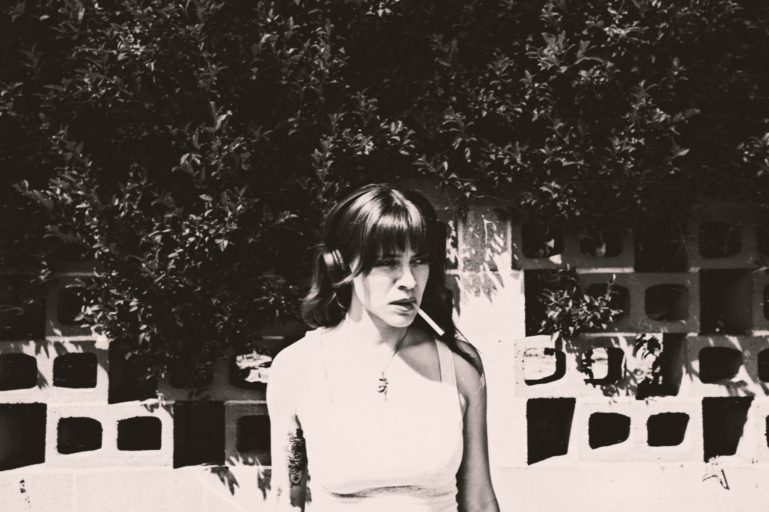 Black and white photo of a woman with long hair and bangs standing outdoors, trees and a wall in background.