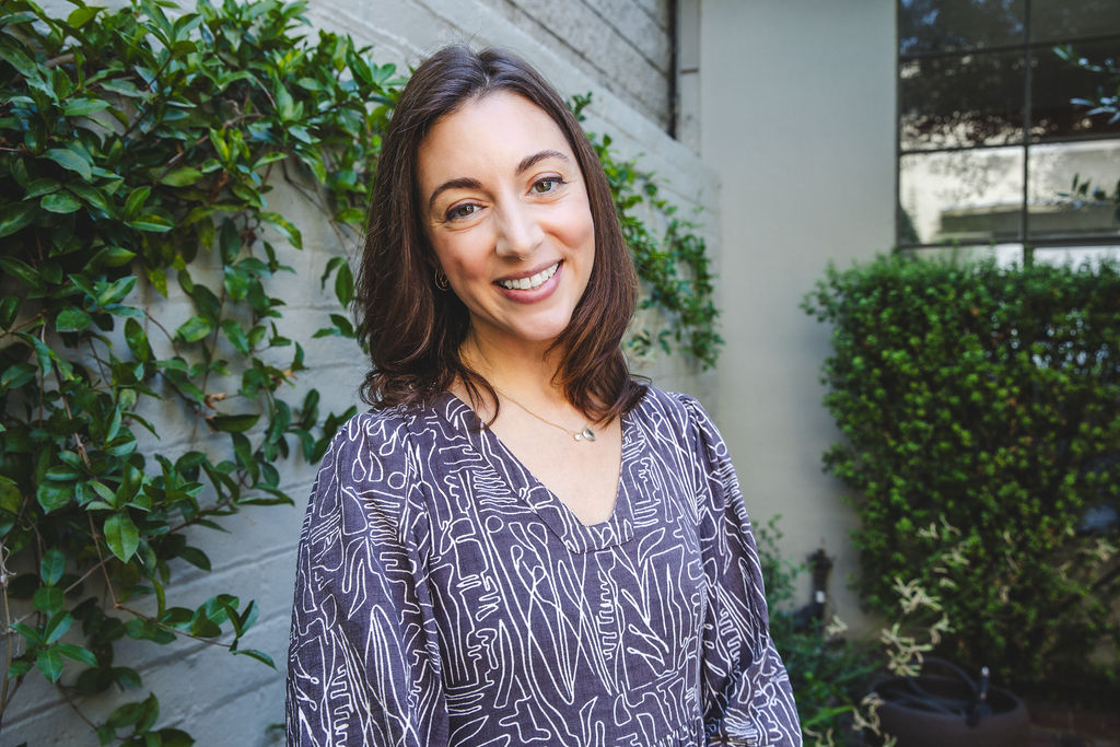 Smiling woman with shoulder-length dark hair outdoors, surrounded by green plants and a building wall.