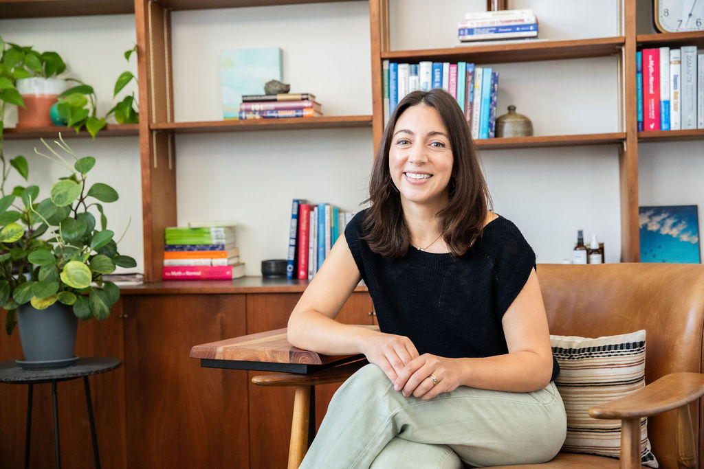 Smiling woman sitting at a wooden table in a room with bookshelves and a potted plant.