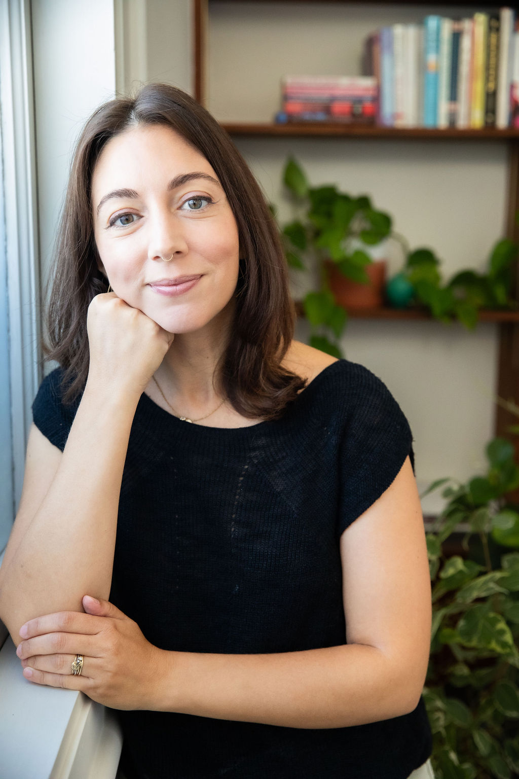 Young woman with shoulder-length brown hair, smiling, resting her chin on her hand, in a room with books and plants