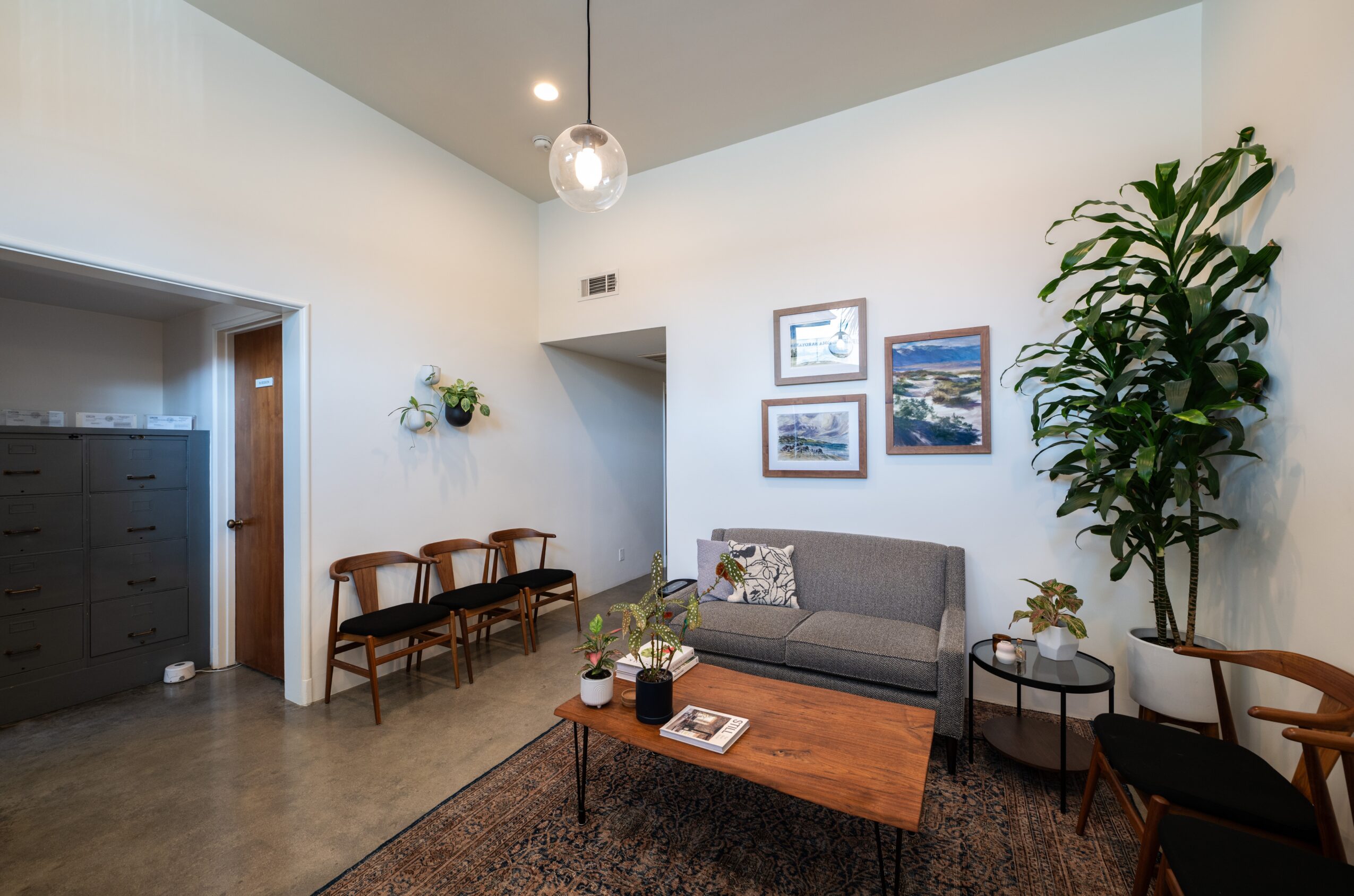 Living room with sofa, wooden coffee table, chairs, wall art, and a tall plant, illuminated by ceiling light.