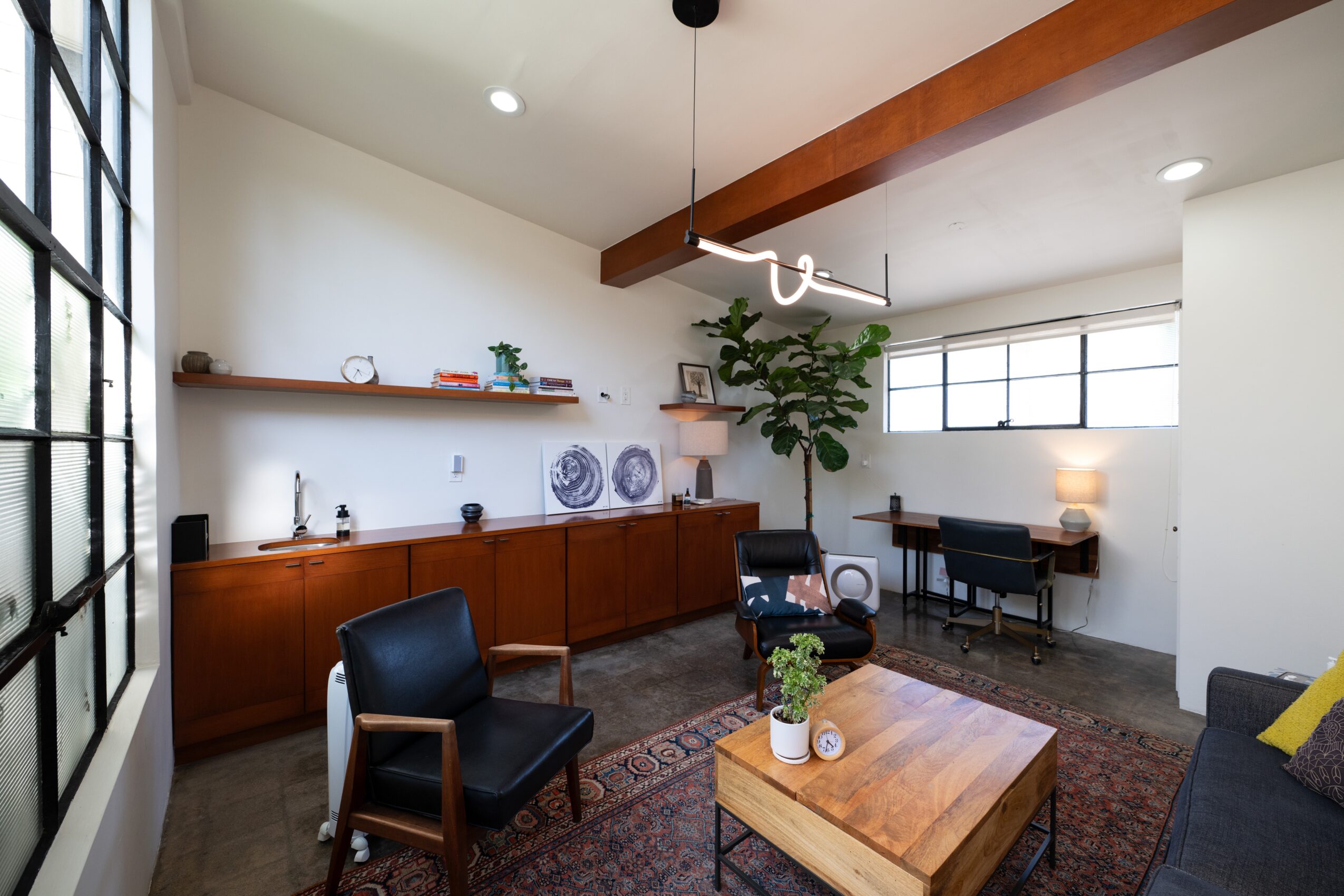 Living room with wooden cabinet, two black chairs, a coffee table with a plant, and a desk with a lamp, window, and ceiling light.