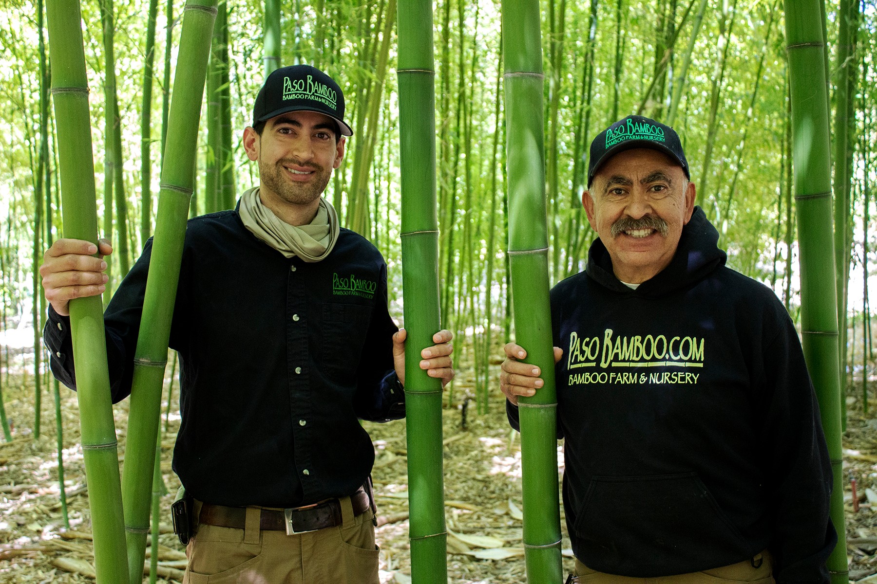 Two men standing among green bamboo stalks, smiling, holding bamboo poles, in a bamboo forest.