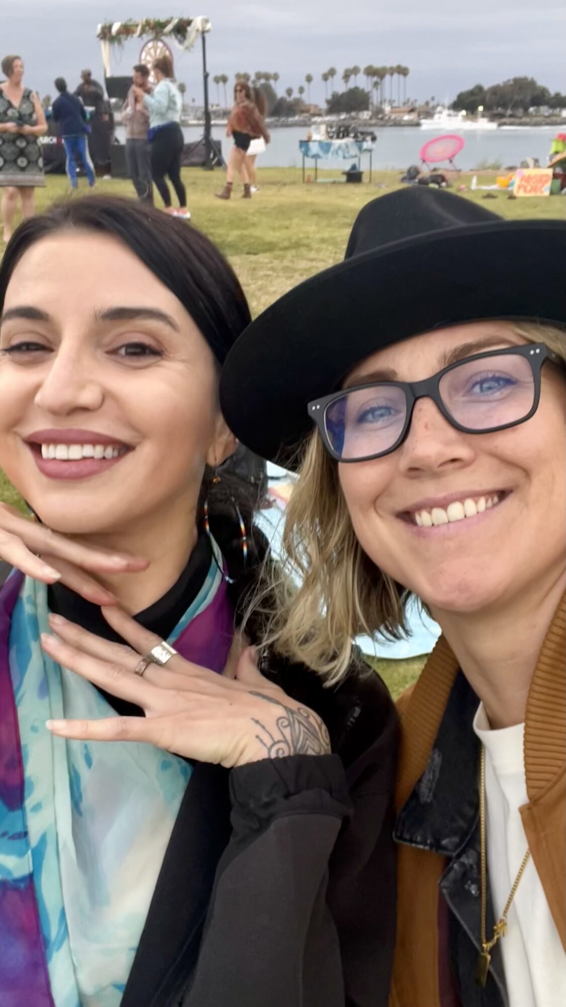Two women smiling outdoors with a park and water in the background, one wearing a hat and glasses.