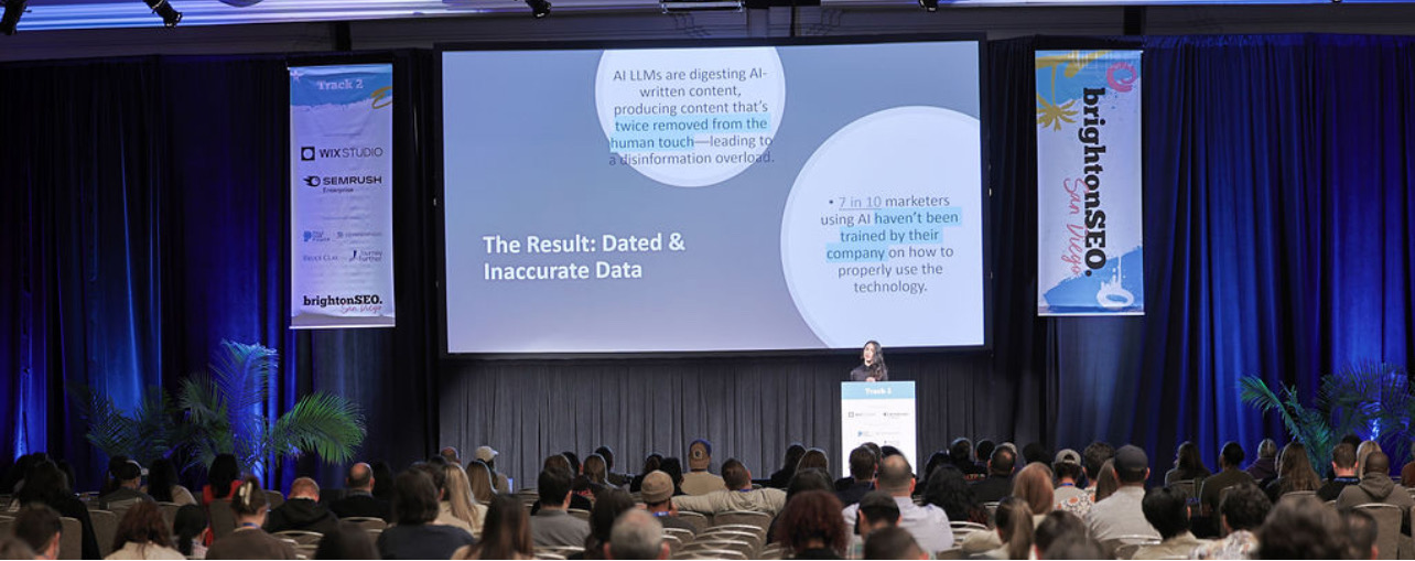 Large conference room with a stage, screen, and audience attending a presentation, with blue curtains and banners.