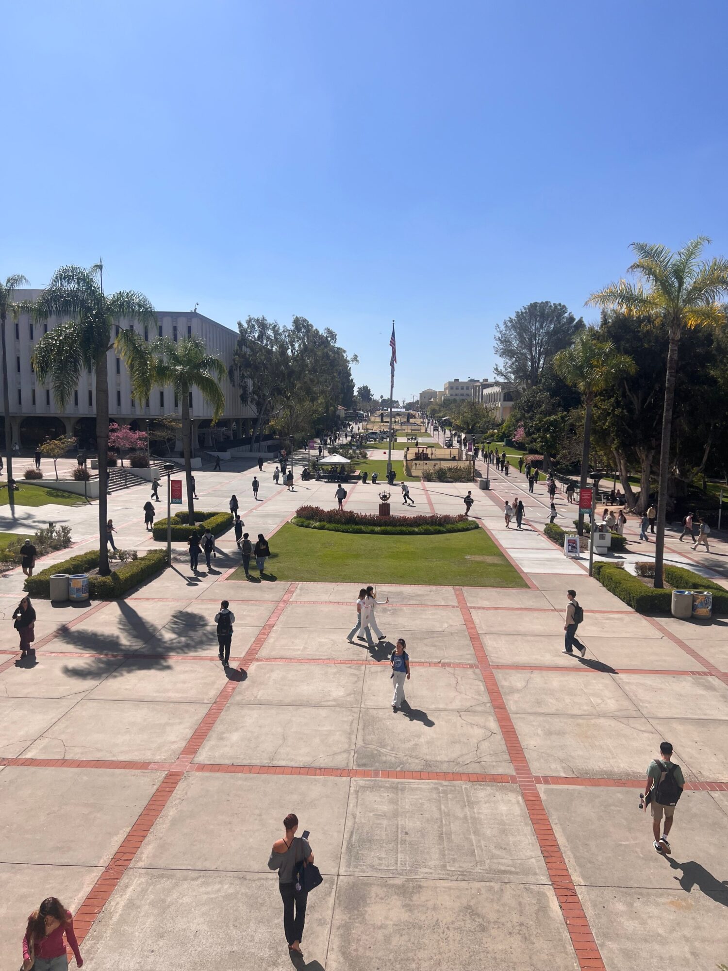 People walking on a sidewalk with trees and buildings in the background, sunny day, shadows cast on the ground.