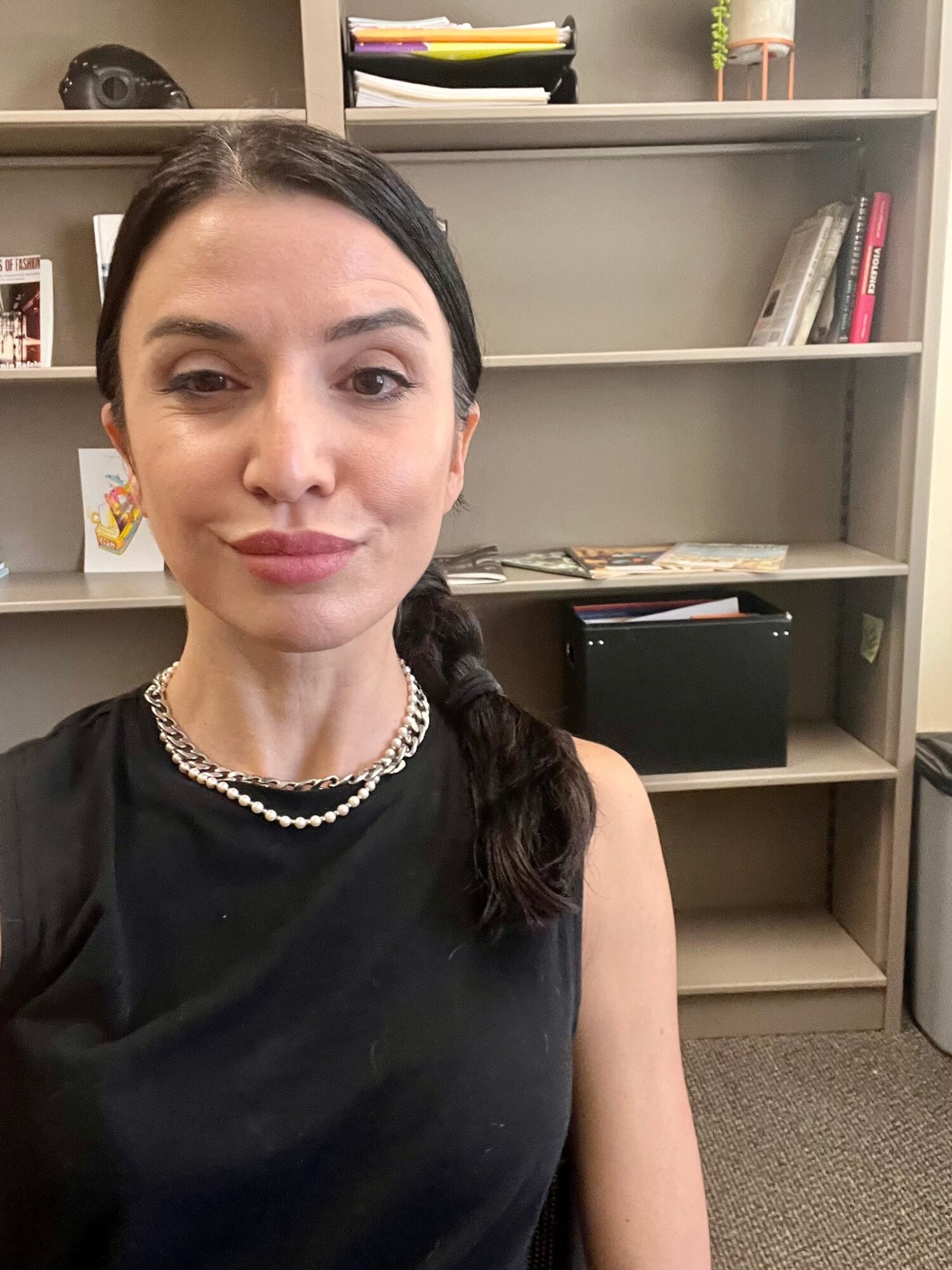 Woman with dark hair in a braid, wearing a black top and pearl necklace, in an office with shelves behind her.