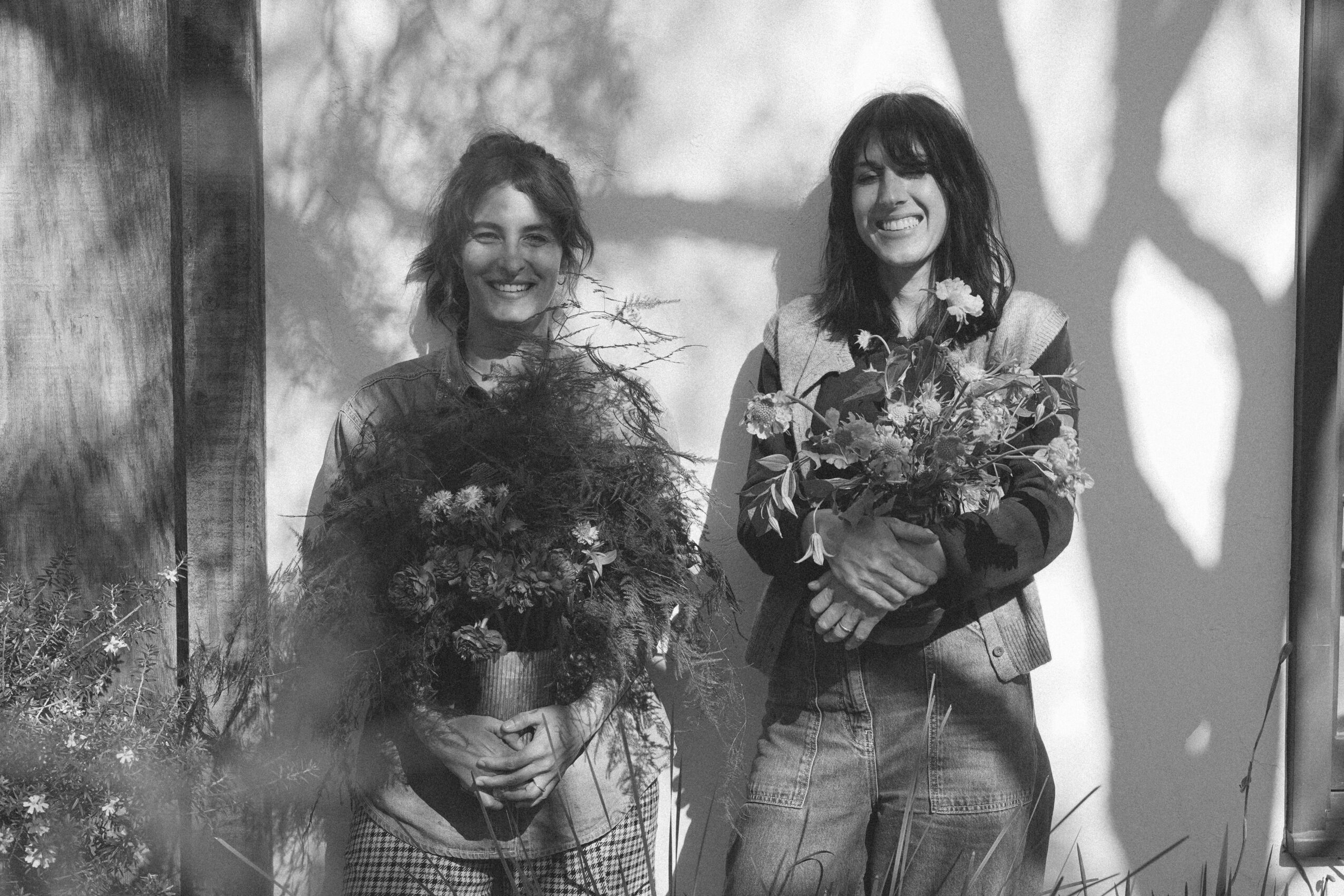 Two women holding large bouquets of flowers, smiling, standing outdoors with trees in the background, black and white photo.