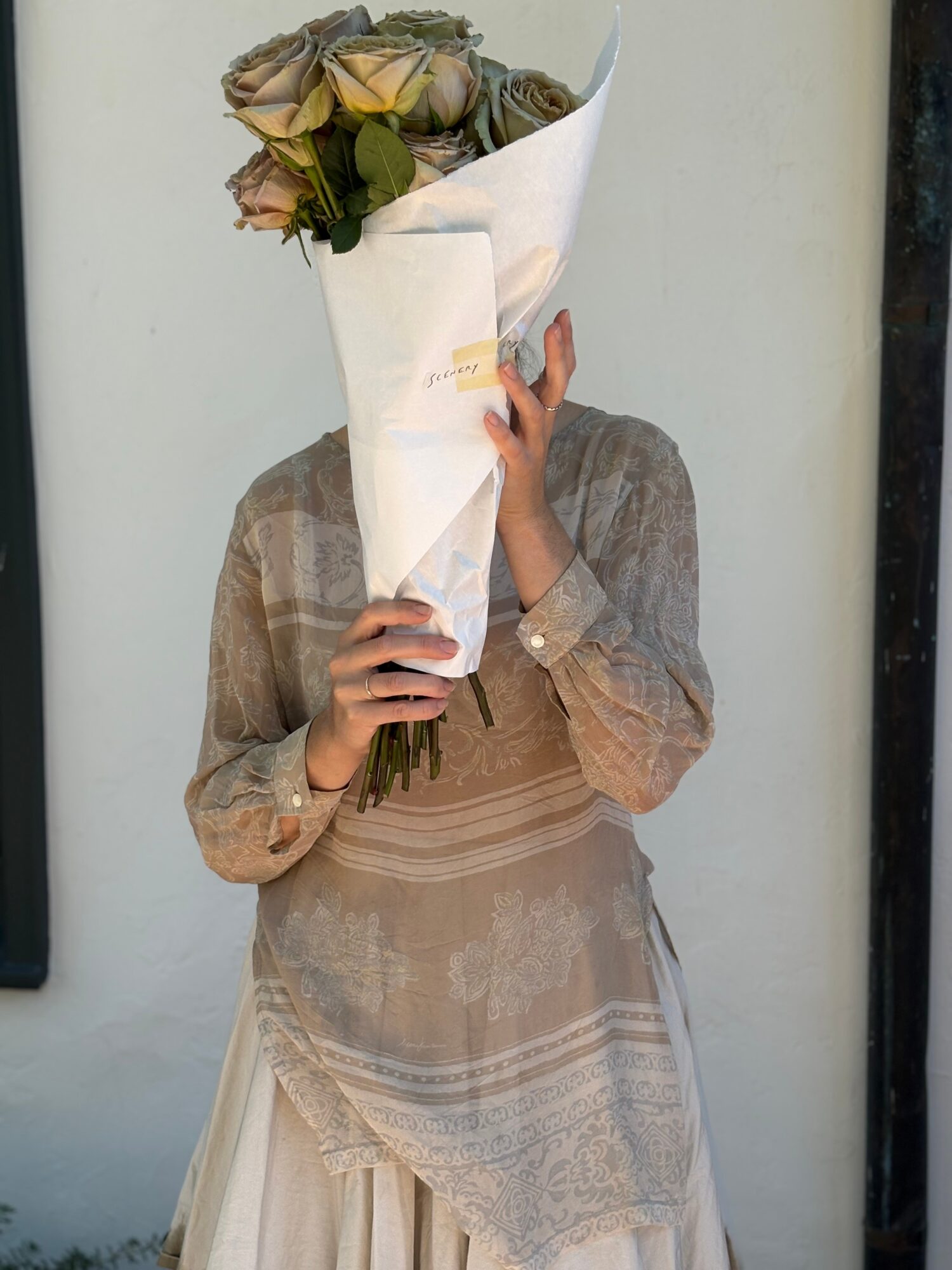 Person holding a bouquet of roses wrapped in white paper, face obscured, wearing a beige dress with lace details.