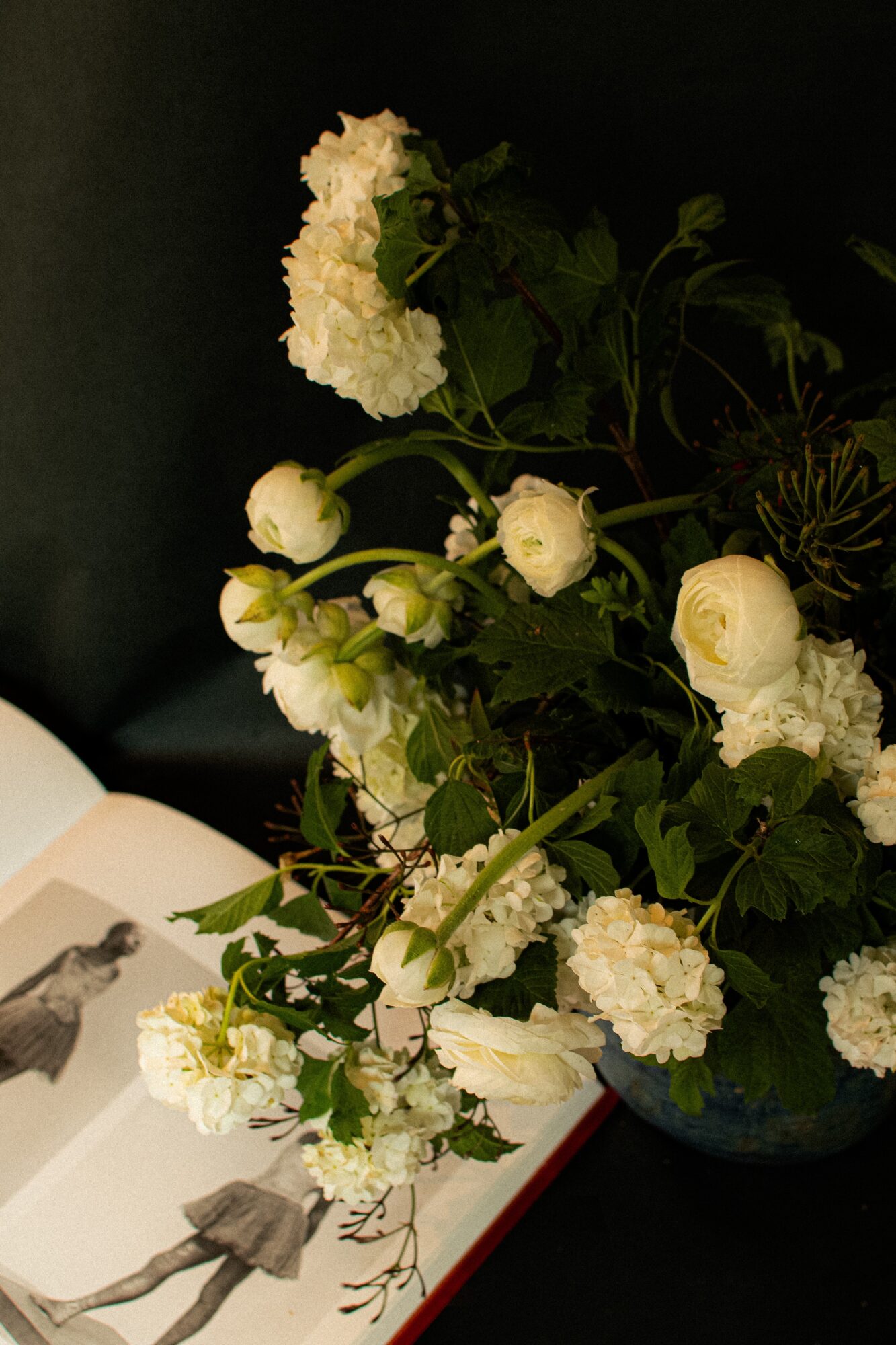 White flowers and green leaves in a dark vase, with an open book nearby, against a dark background.