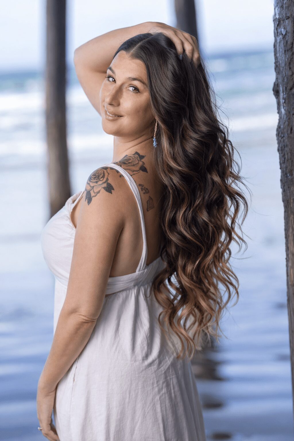 Young woman with long wavy hair, smiling, standing outdoors near water, wearing a white sleeveless dress, with a tattoo on her shoulder.