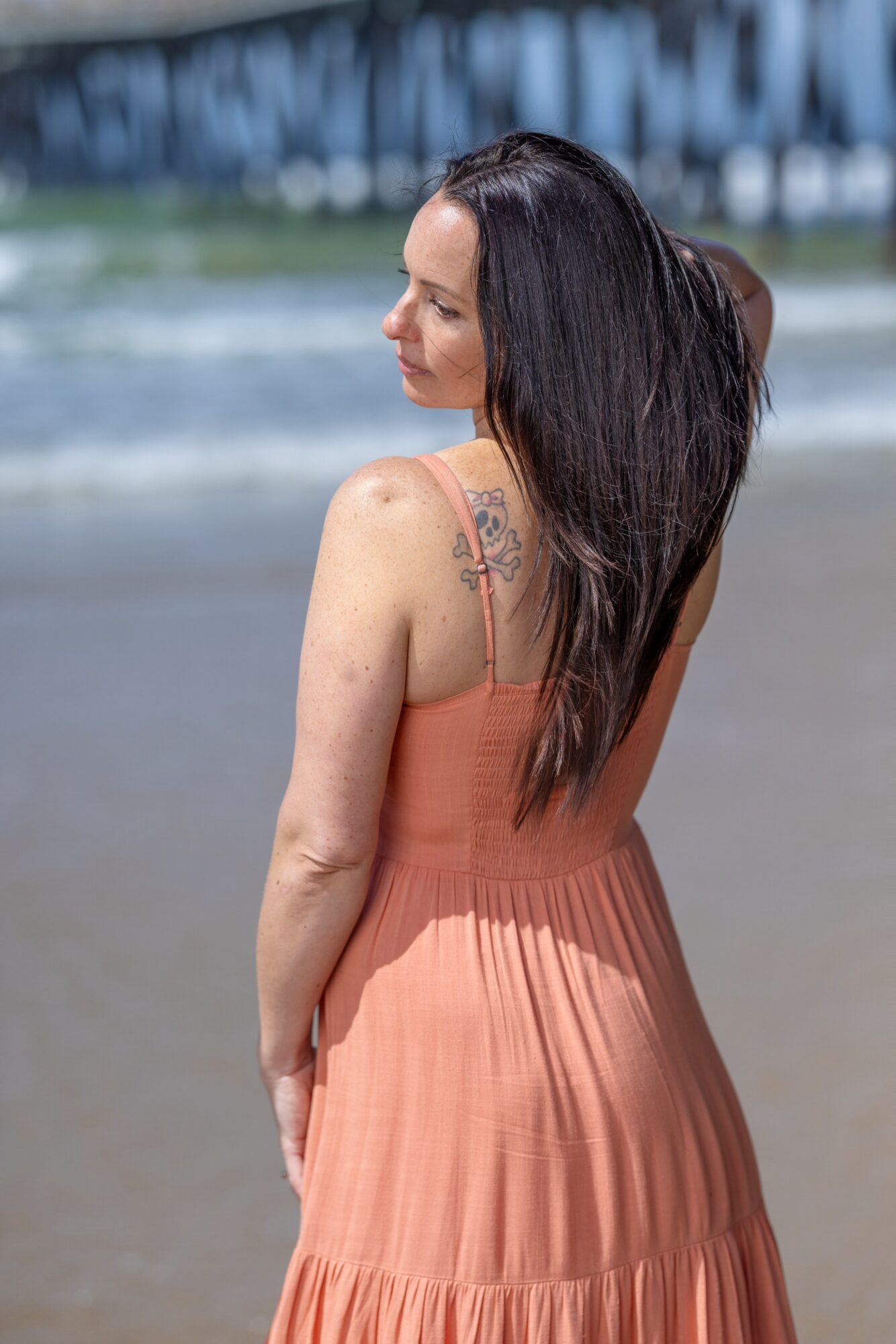 Woman with long dark hair in a peach dress standing on a beach, facing sideways with her eyes closed.