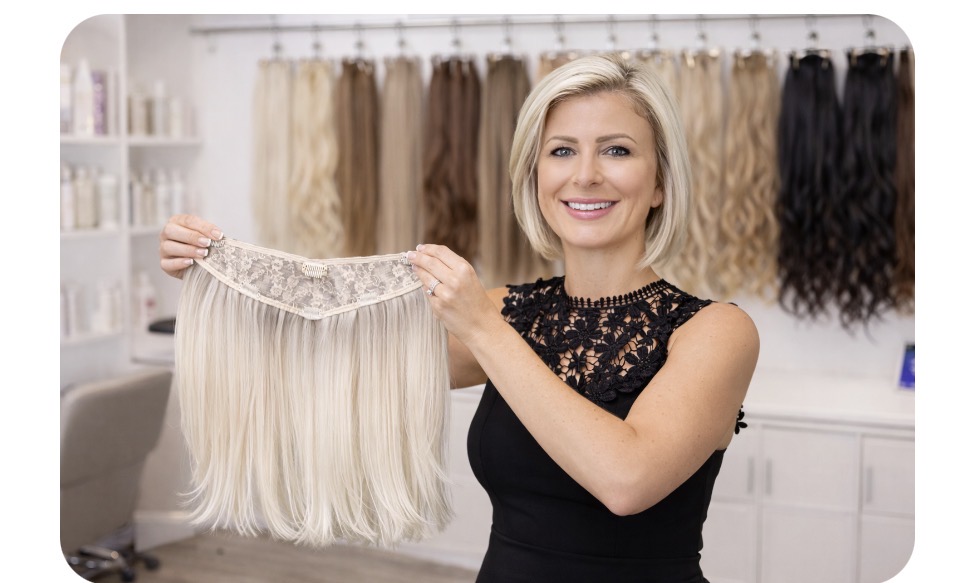 Woman holding a blonde hair extension piece in a hair store, smiling, with hair extensions on shelves behind her.