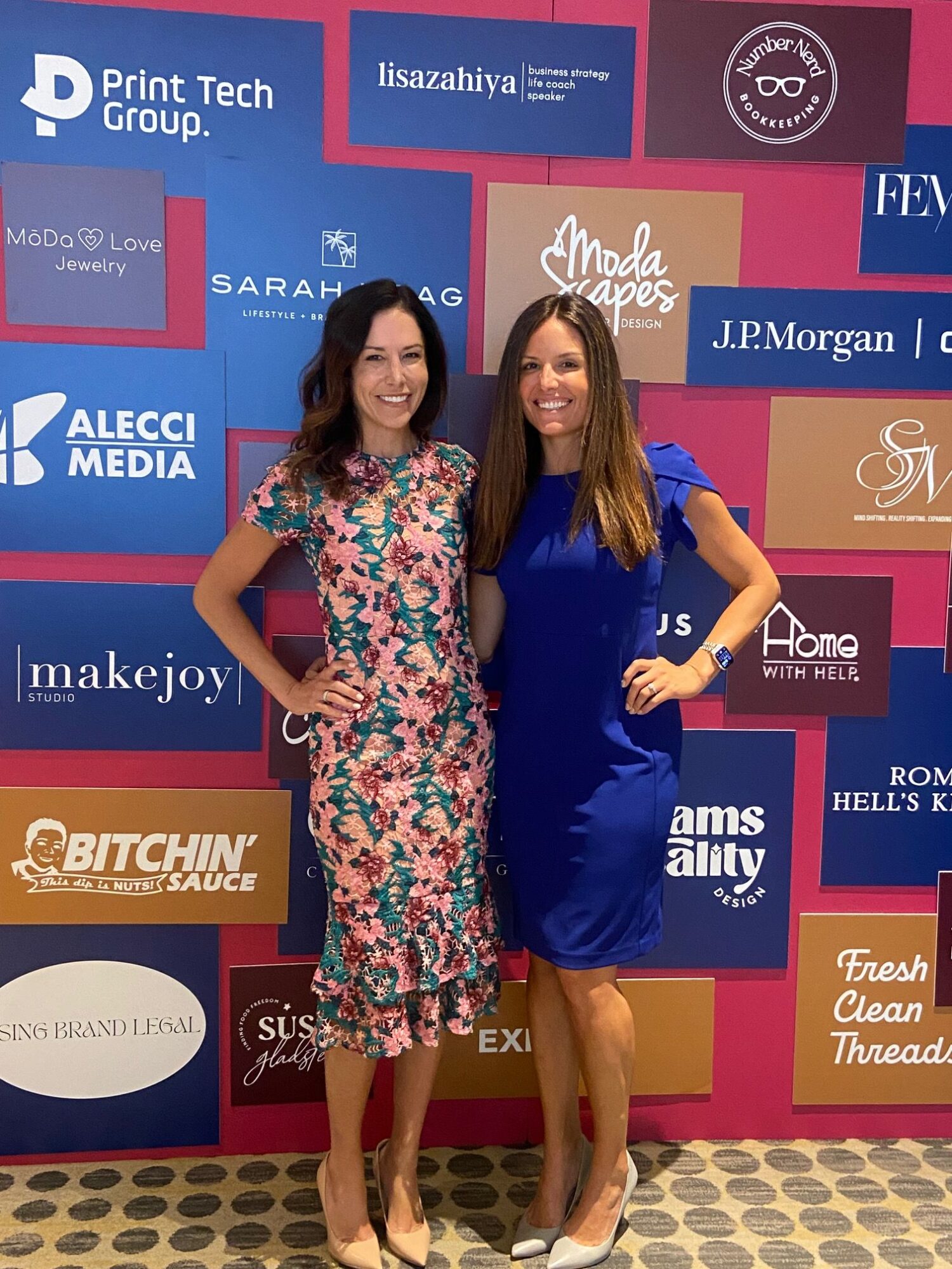 Two women standing in front of a colorful backdrop with various logos, one in a blue dress and the other in a floral dress.