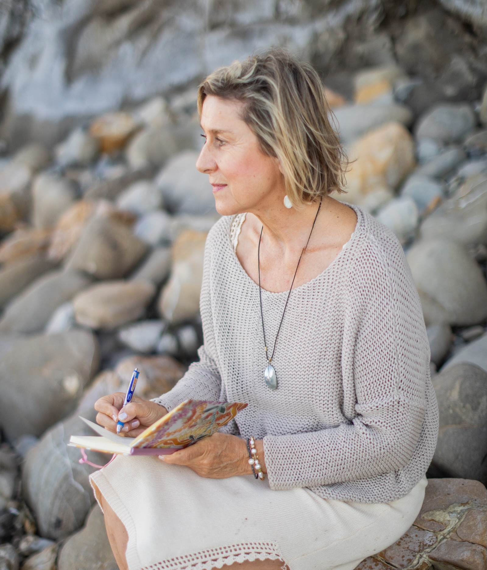Woman sitting on rocks, reading a book, wearing a light sweater and necklace, with a rocky background.