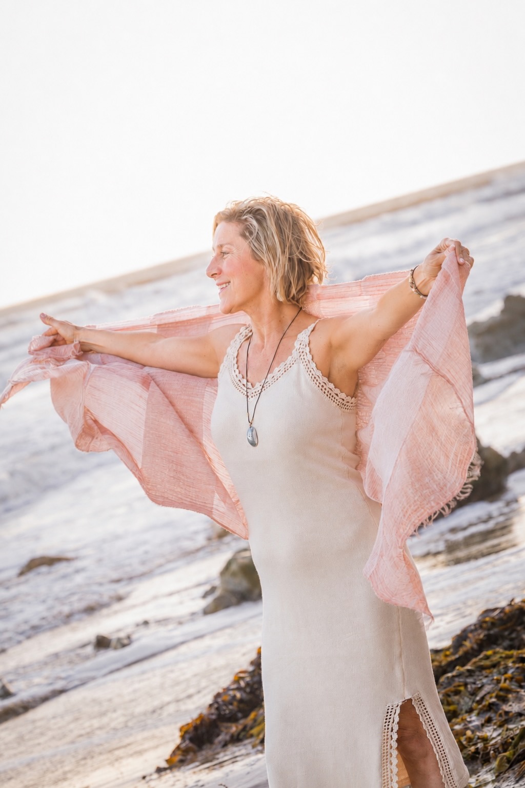 Woman standing on rocks at the beach with arms outstretched, holding a pink shawl, smiling, ocean in background