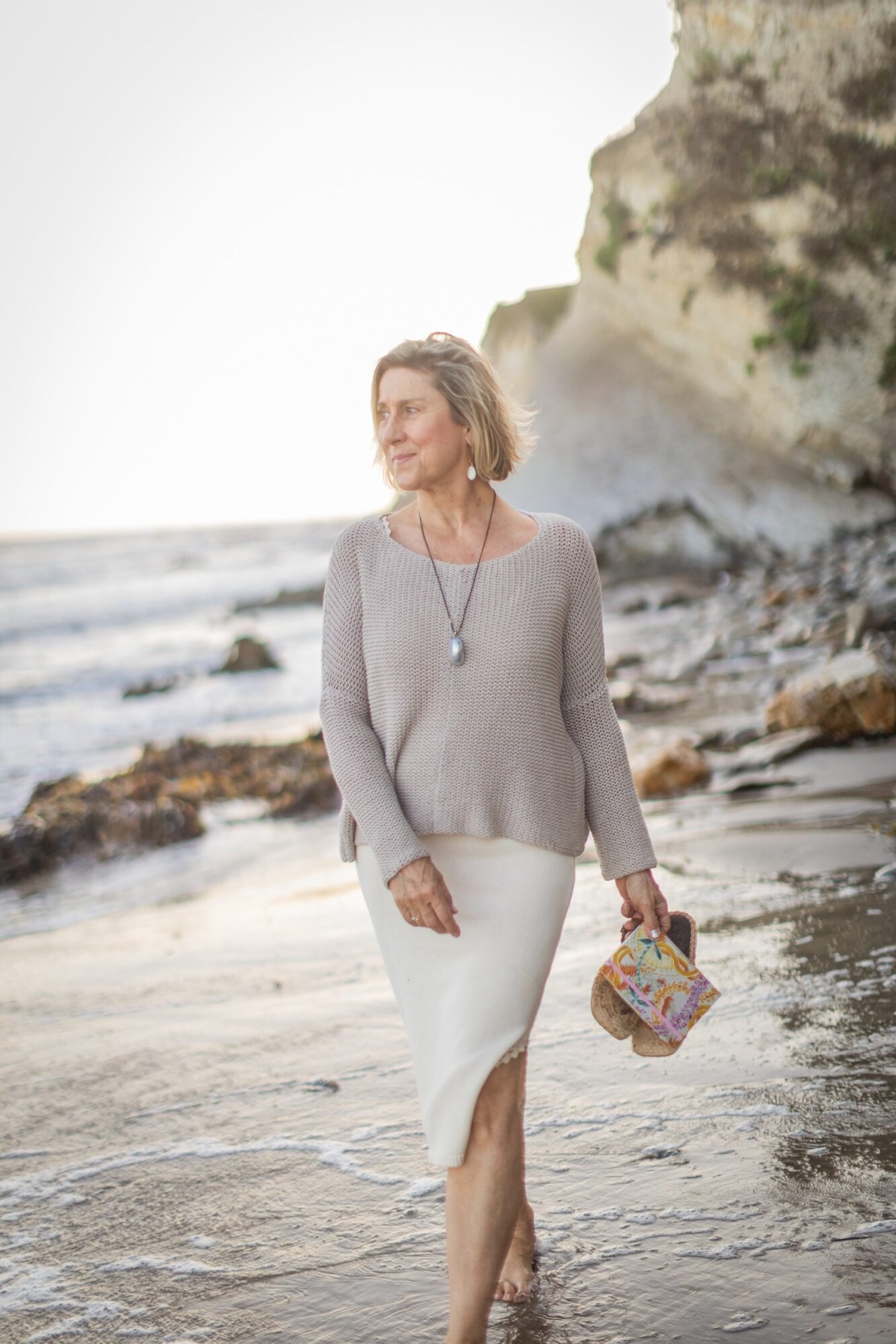 Woman walking on a beach with cliffs in the background, holding a colorful bag, wearing a gray sweater and white skirt.