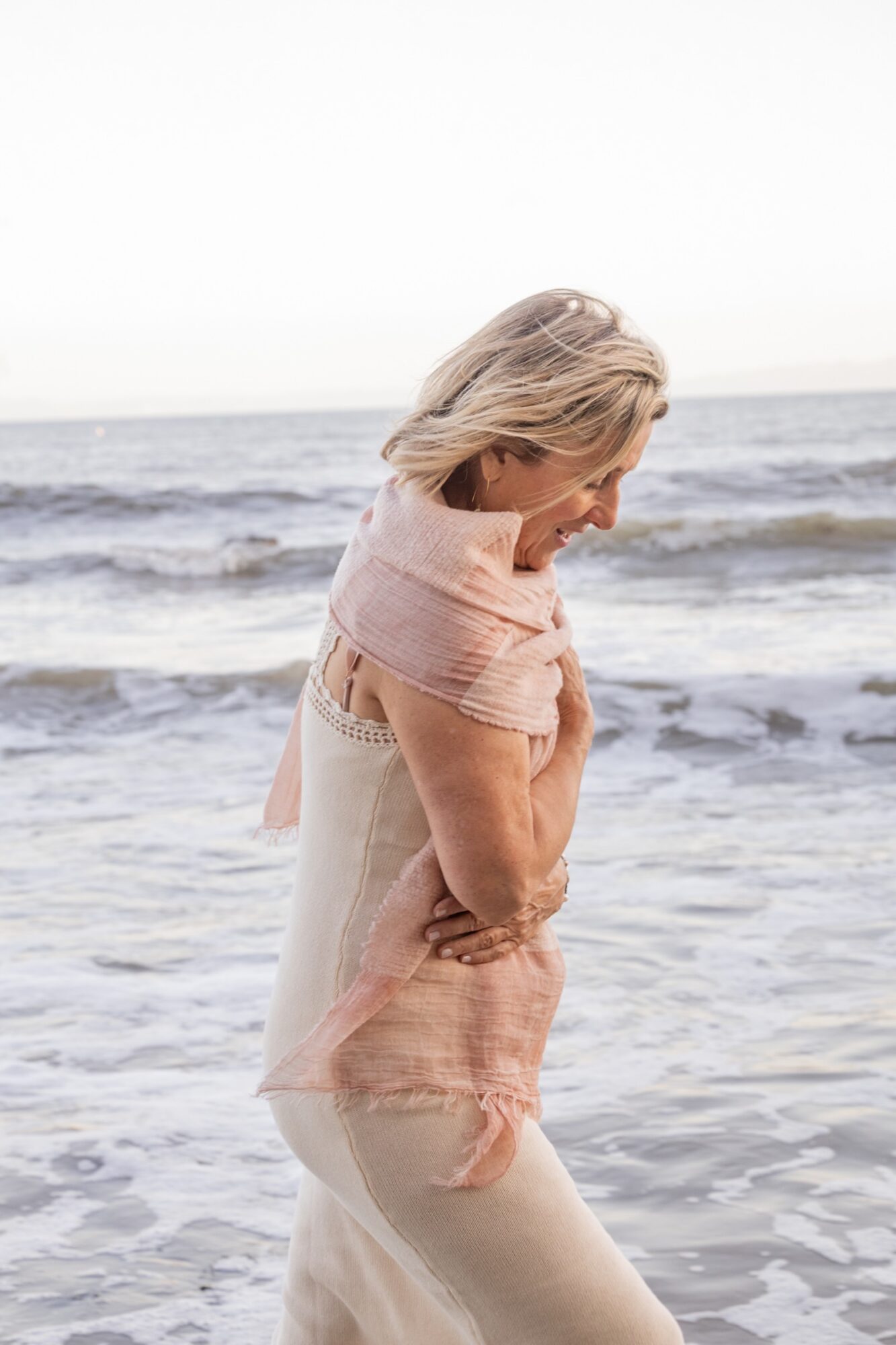 Woman standing on the beach with waves in the background, looking down, wearing light-colored clothing and a pink shawl.