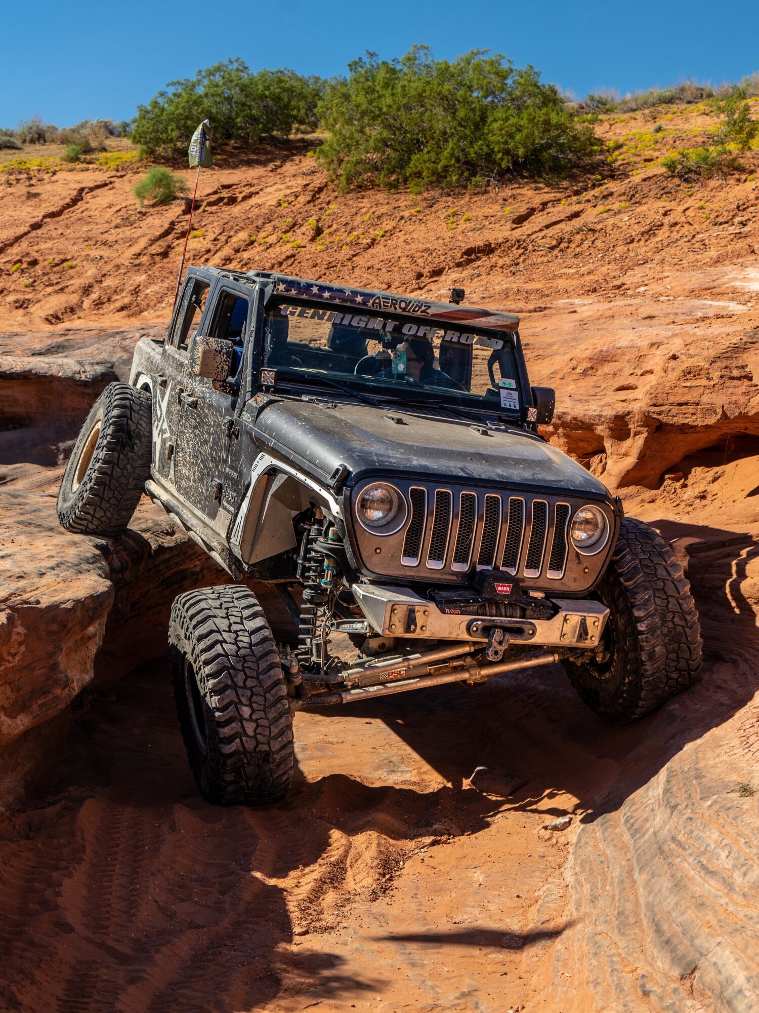 Off-road vehicle navigating rocky terrain with large tires and a rugged exterior, desert landscape with bushes and blue sky in background.