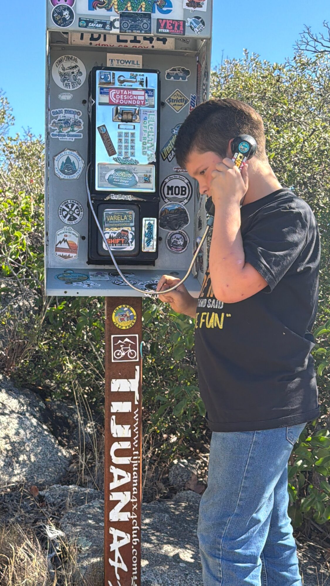Boy using a public payphone with headphones, outdoors, trees, and rocks in background.