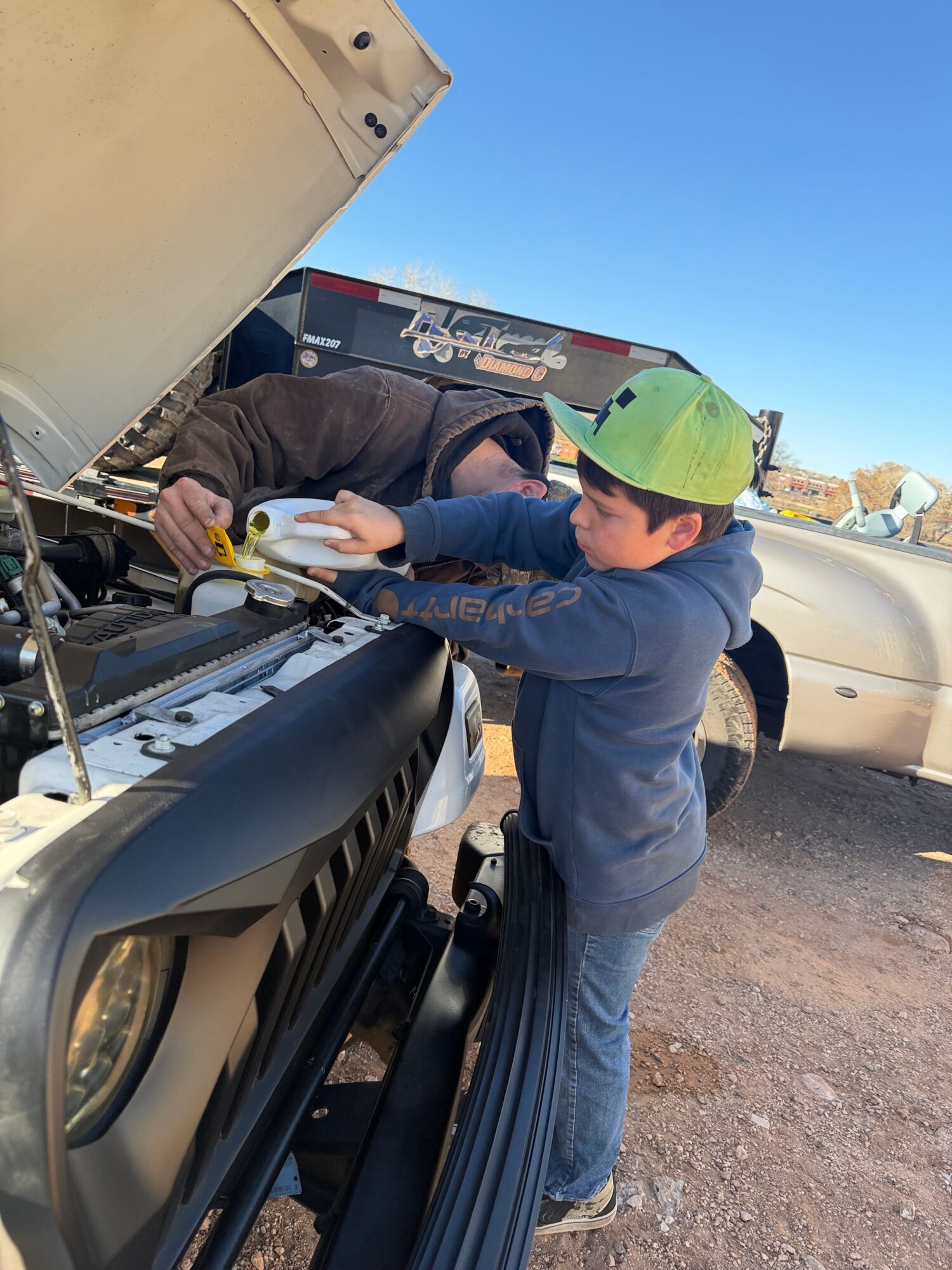 Two people working on a motorcycle, one wearing a green cap and blue jacket, outdoors on a sunny day.
