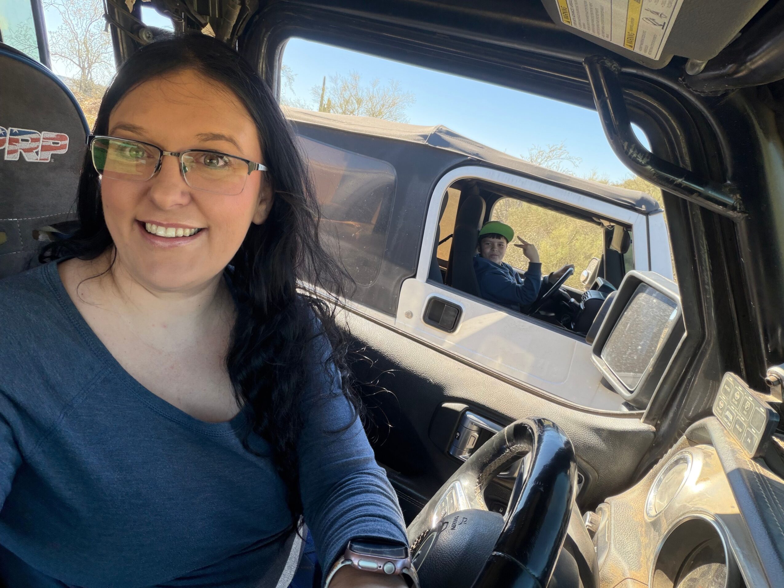 Woman with glasses smiling inside vehicle, dashboard and side window visible, sunny day outside.