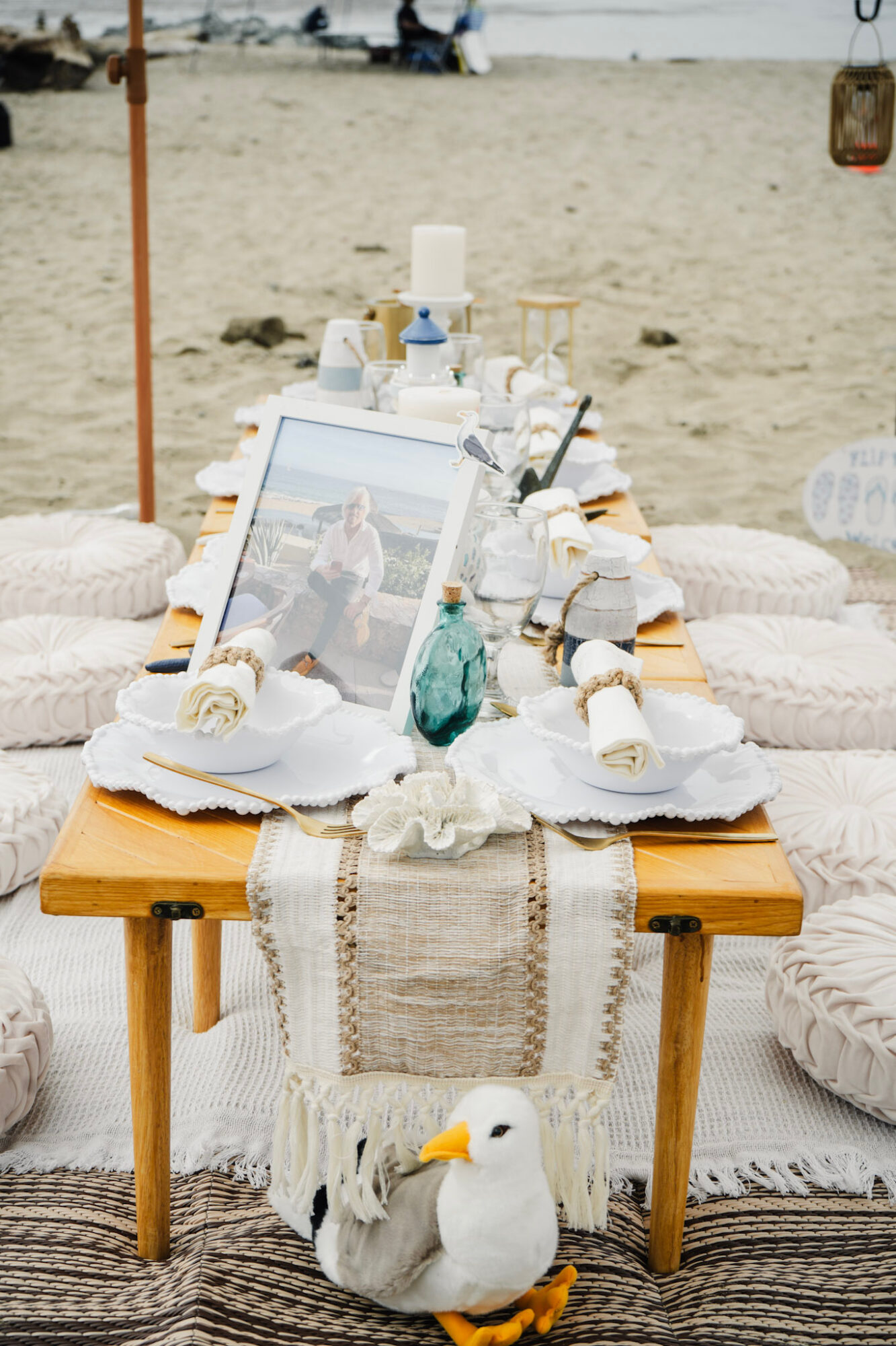 Beach table with framed photo, candles, and decorative items, surrounded by white cushions and a seagull figure, on sandy beach.